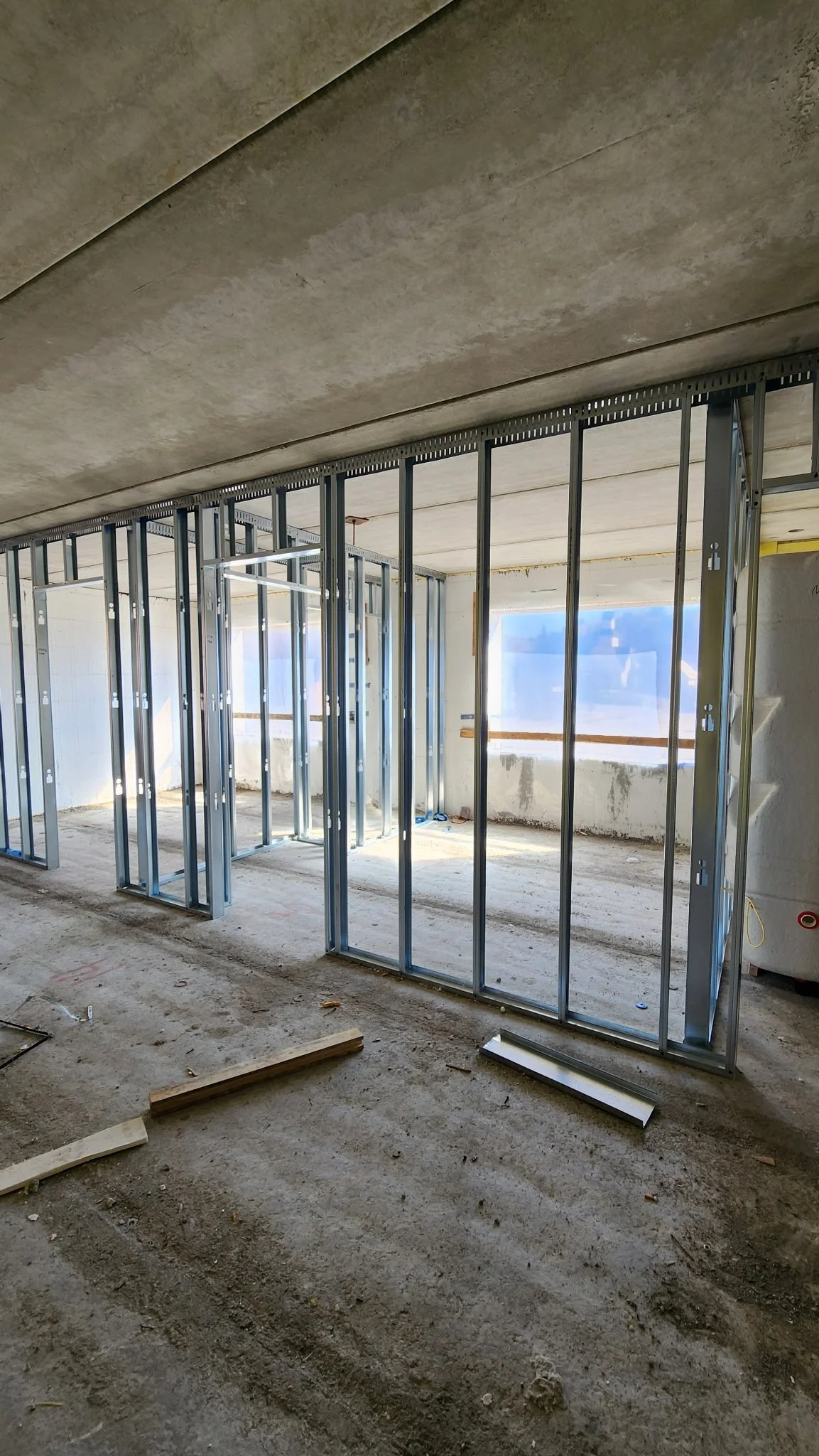 Interior view of a building under construction, showing metal framing for walls, unfinished floors, and large windows with sunlight coming through.