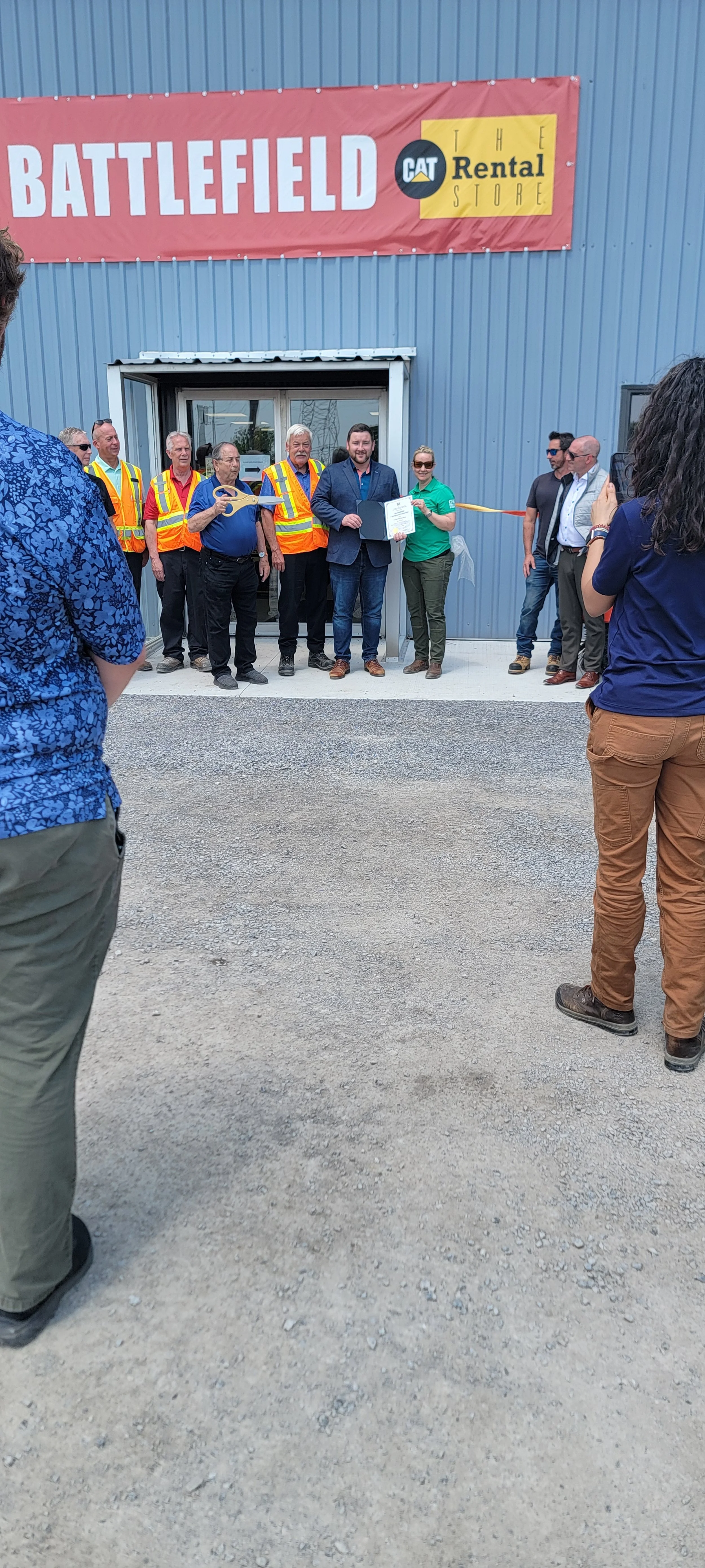 A group of people gathered outside a building with a sign that reads 'Battlefield CAT Rental Store'. Some individuals are wearing safety vests, and one person is holding a large pair of scissors, suggesting a ribbon-cutting ceremony.