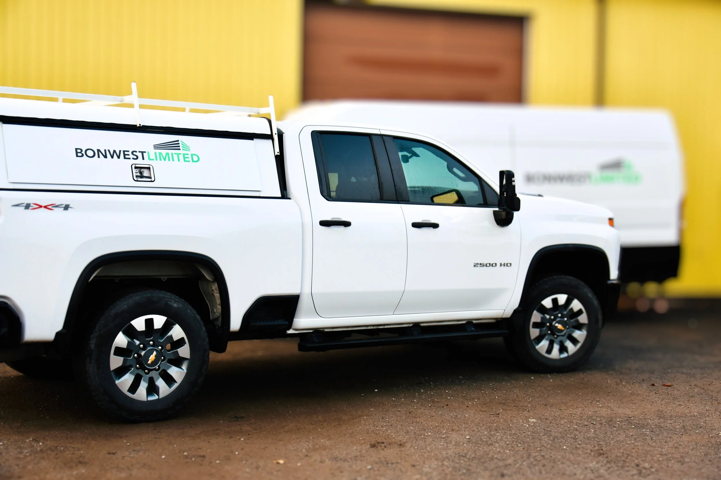 White Chevrolet Silverado pickup truck with a service bed, parked on a dirt surface, with a yellow building background. The vehicle has black side mirrors and Chevrolet logo on the wheels, with branding for Bonwest Limited on the service bed and a white van in the background.
