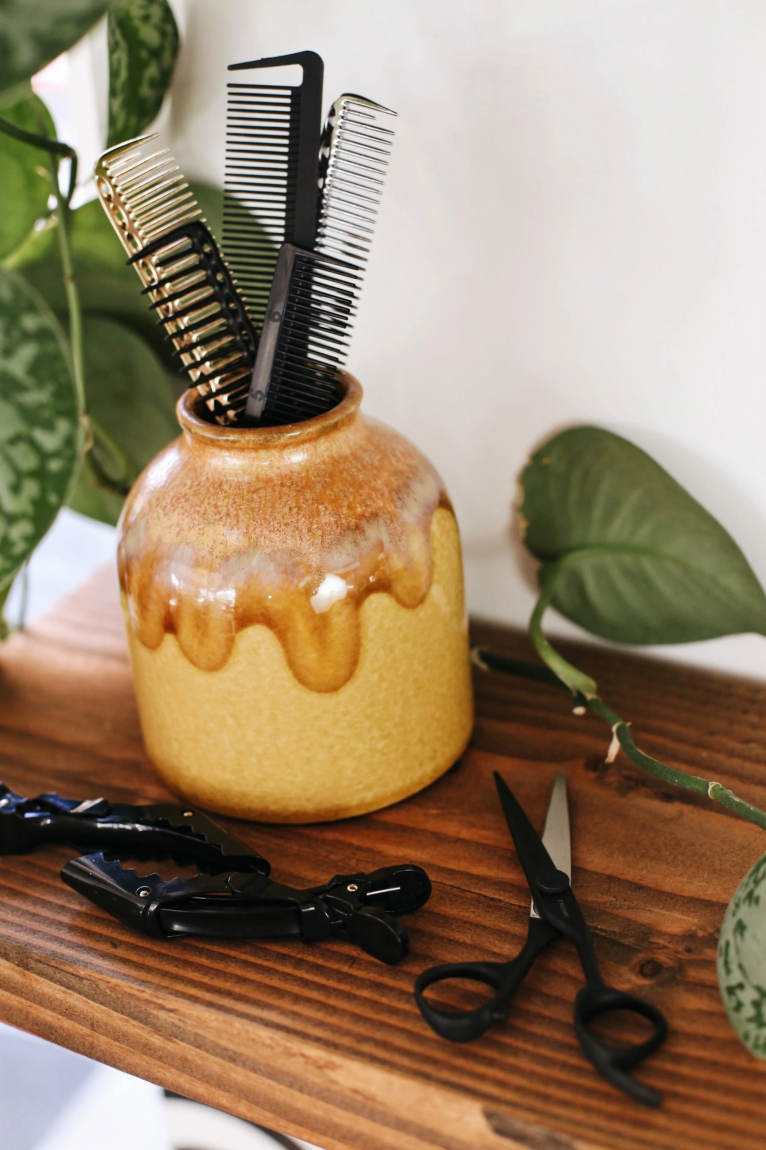 A small, yellow ceramic vase holding black and silver hair combs, with black scissors and a black hair clip on a wooden table, surrounded by green leafy plants.