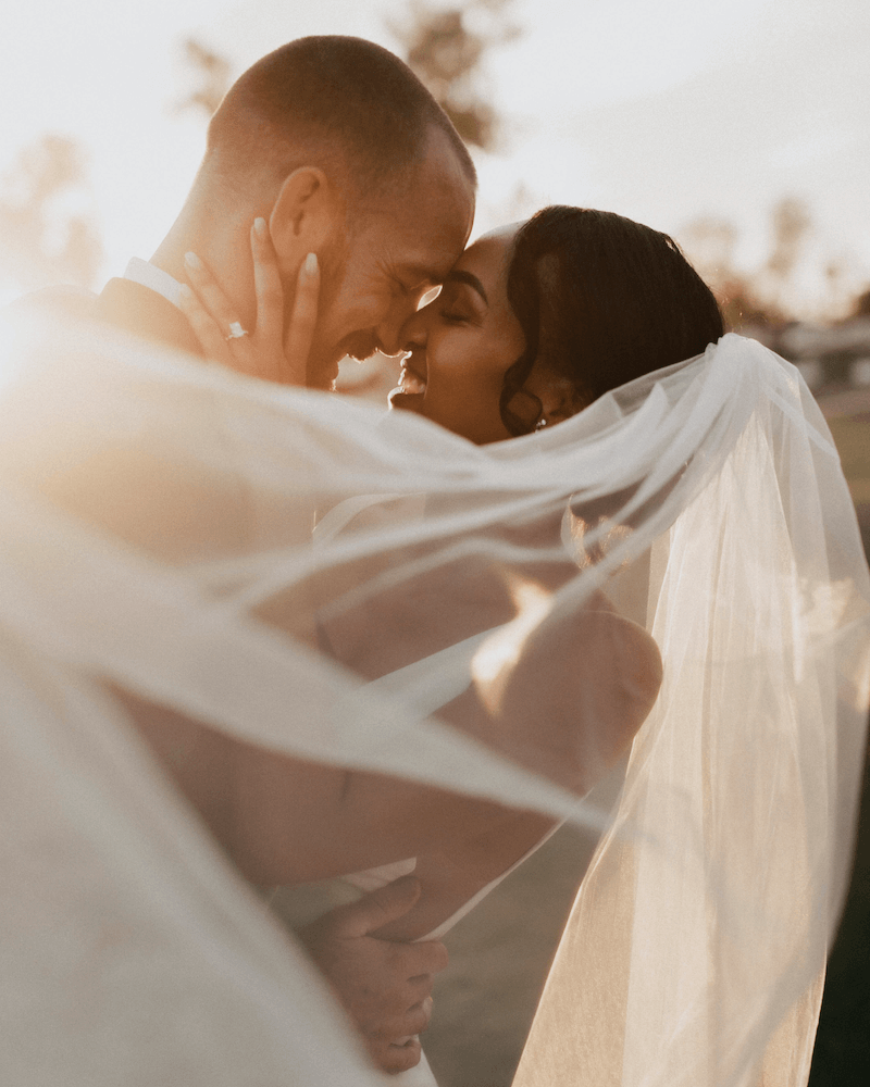 A close-up of a bride and groom embracing, with their foreheads touching, smiling and gazing at each other at sunset on their wedding day.
