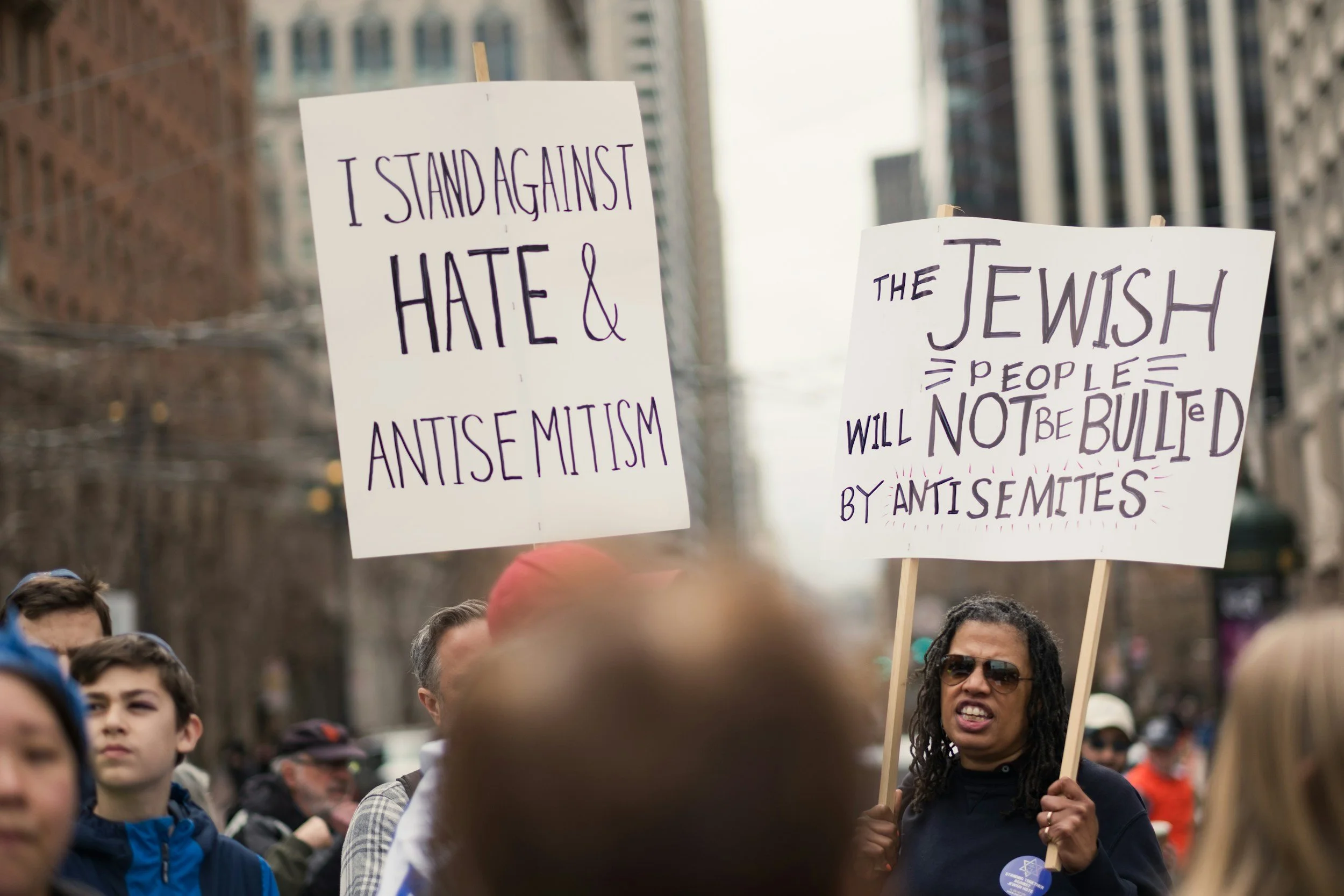 People participating in a protest march holding signs against antisemitism in an urban setting with tall buildings. One sign reads, "I stand against hate & antisemitism," and the other states, "The Jewish people will not be bullied by antisemites."