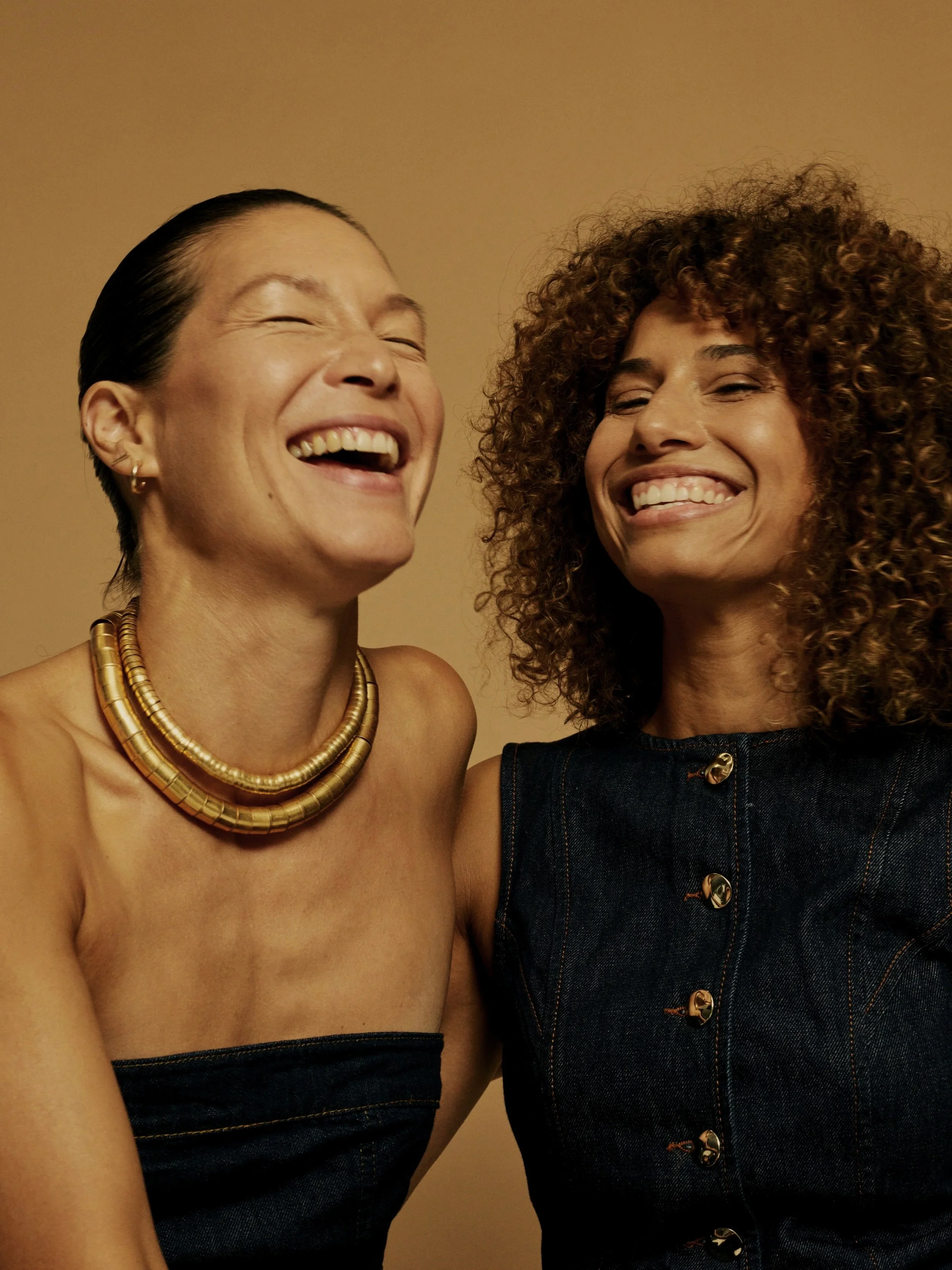 Two women smiling and laughing together, one with short dark hair and gold jewelry, the other with curly hair wearing a denim sleeveless top.