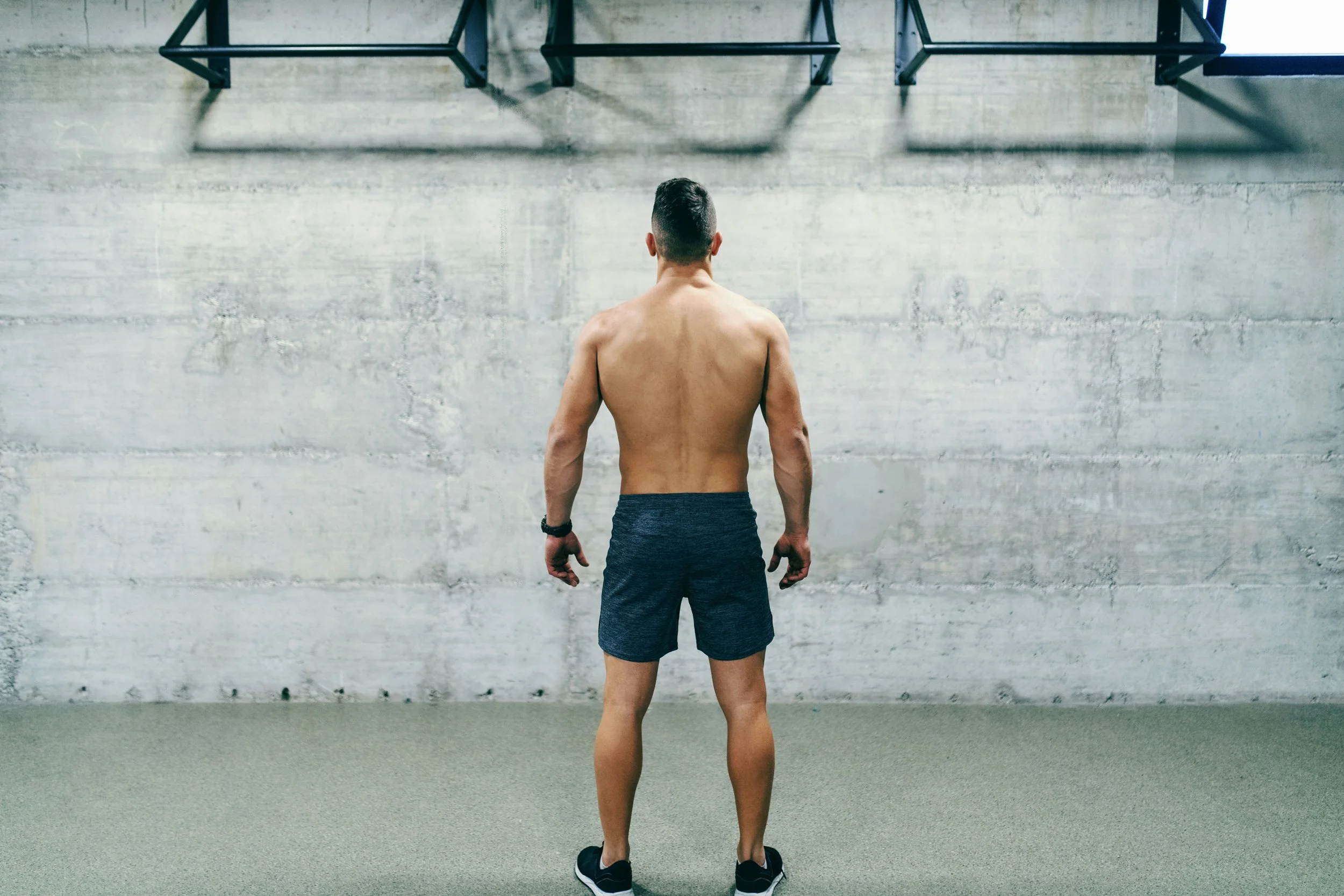 A shirtless man with dark hair, wearing black shorts and sneakers, standing with his back to the camera in an indoor gym or fitness center.