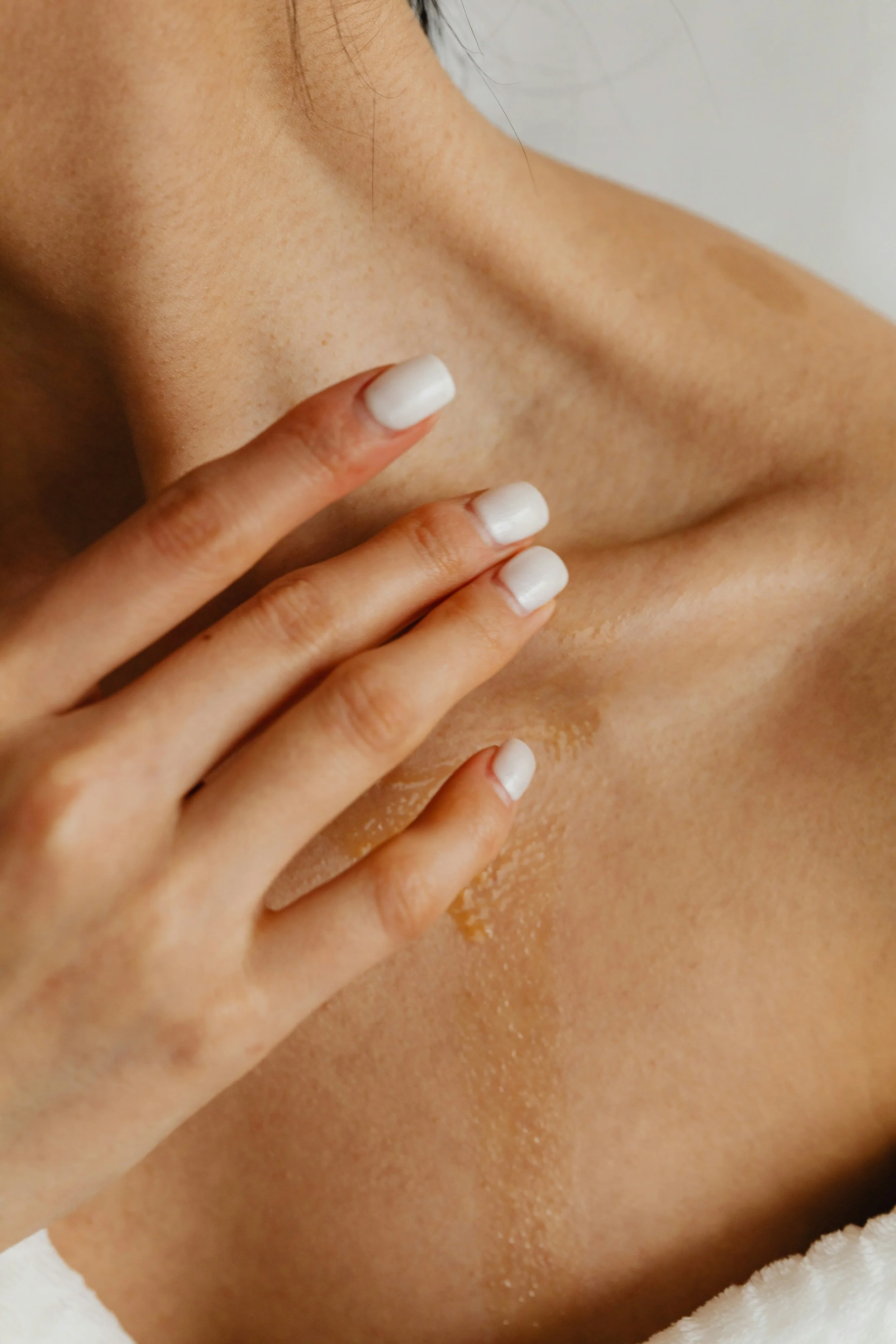 Close-up of a woman's shoulder and collarbone area with hand touching the skin, showing smooth skin and white painted nails.