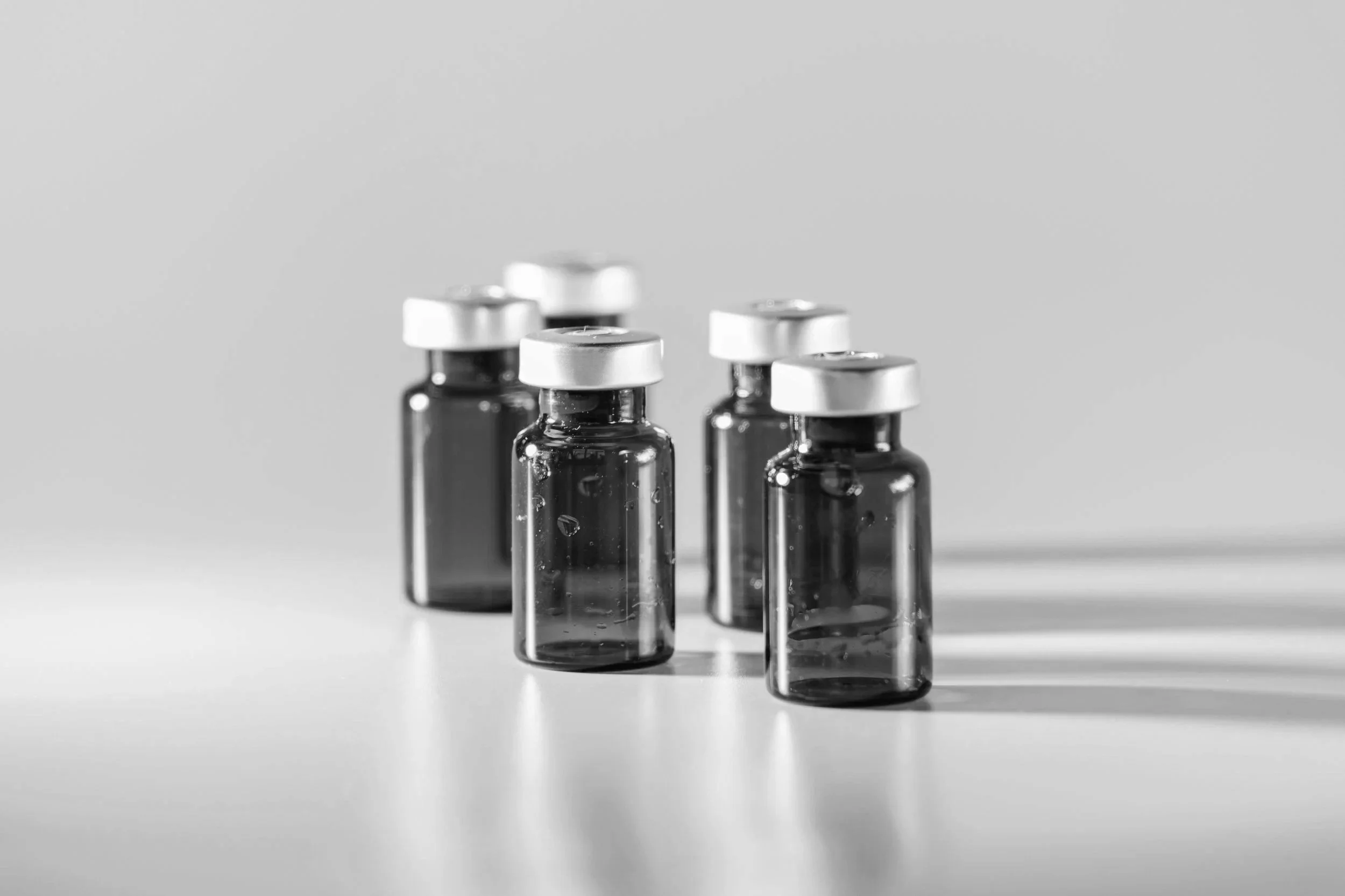 Five small glass vials with silver caps, arranged on a white surface, casting shadows, in black and white.