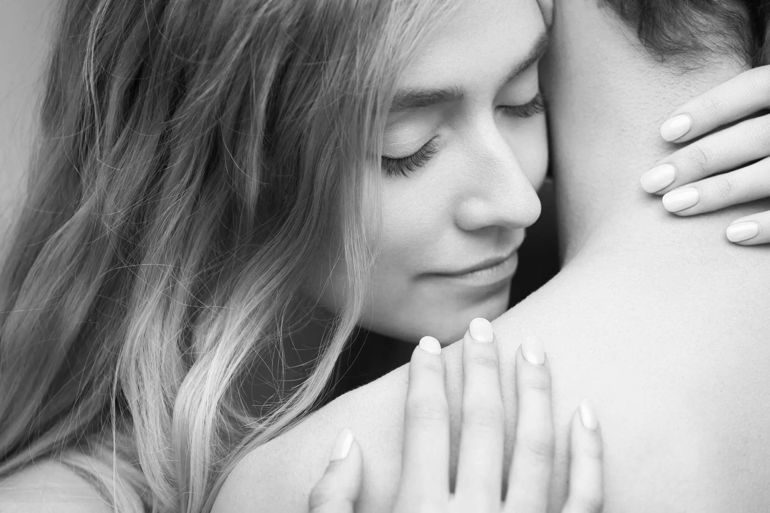 A woman with long wavy hair gently embracing and resting her face against a person with short hair, both with closed eyes, in a tender, intimate moment.