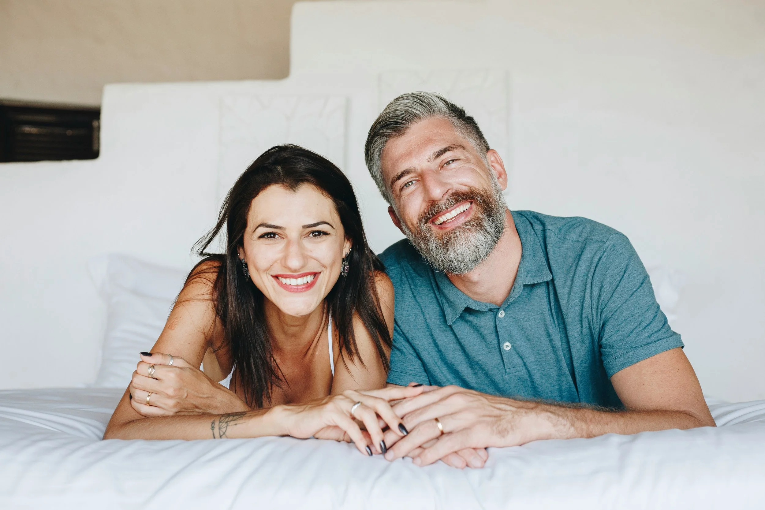 A smiling couple lying on a bed, close together, with the woman on the left and the man on the right, both looking at the camera.