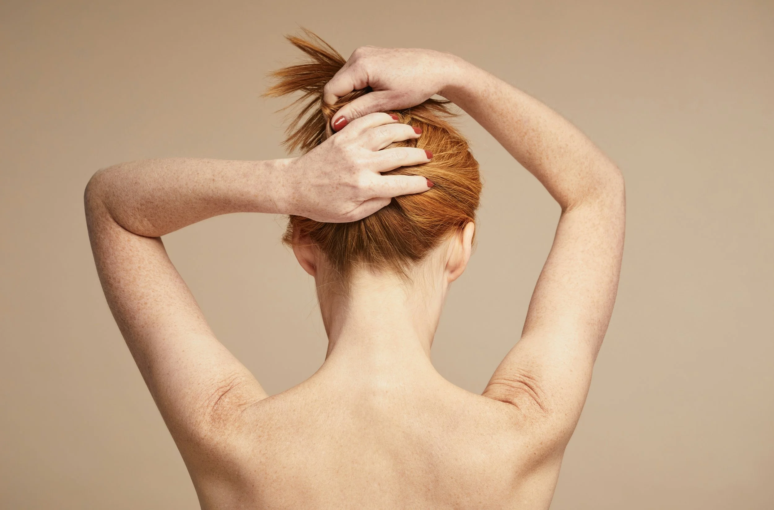Back of a person with short red hair, holding their hair with both hands, posing against a beige background.