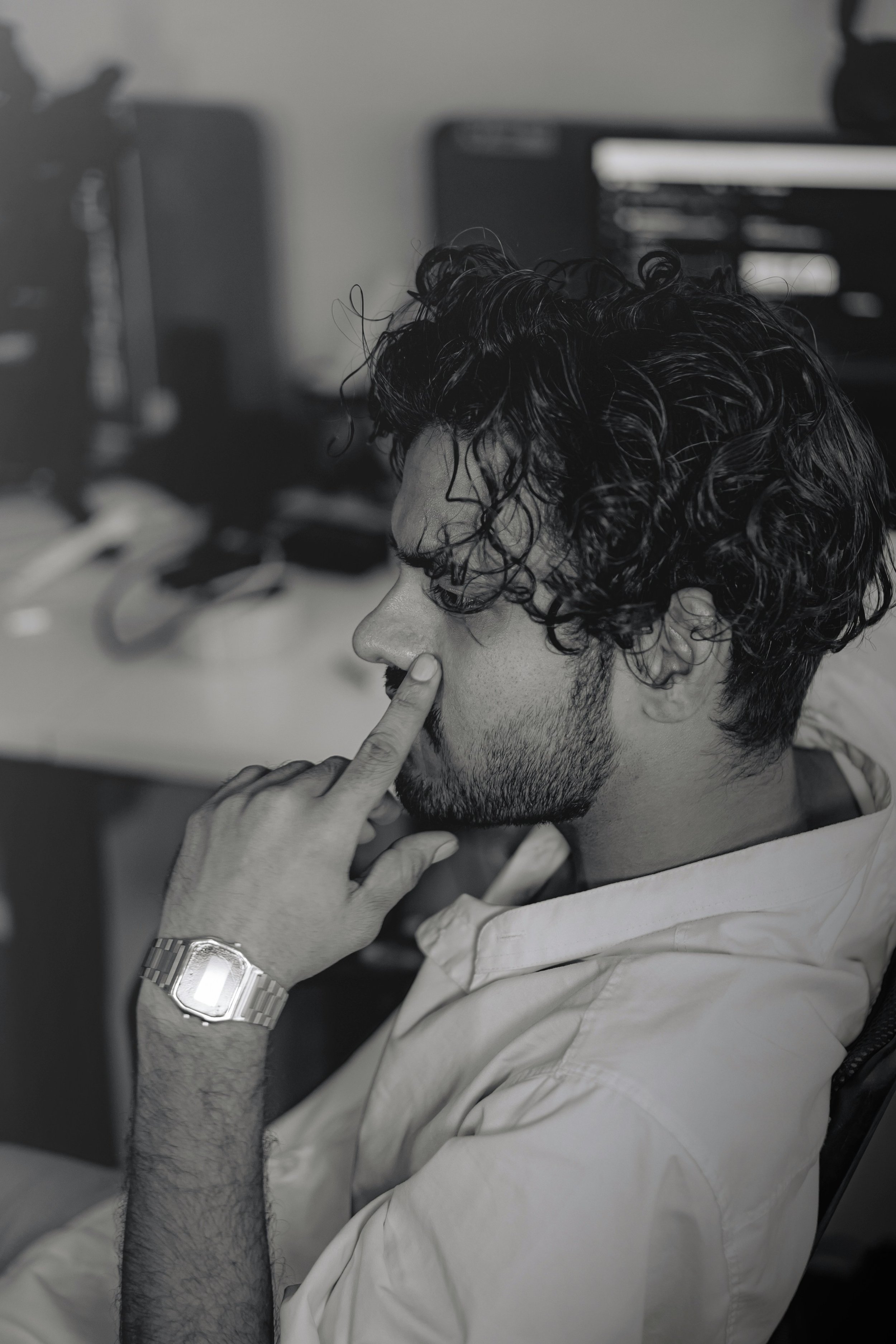 A man with curly dark hair, beard, and mustache, wearing a wristwatch and a light-colored hoodie, is sitting in front of a computer, touching his lips with his index finger, indoors.