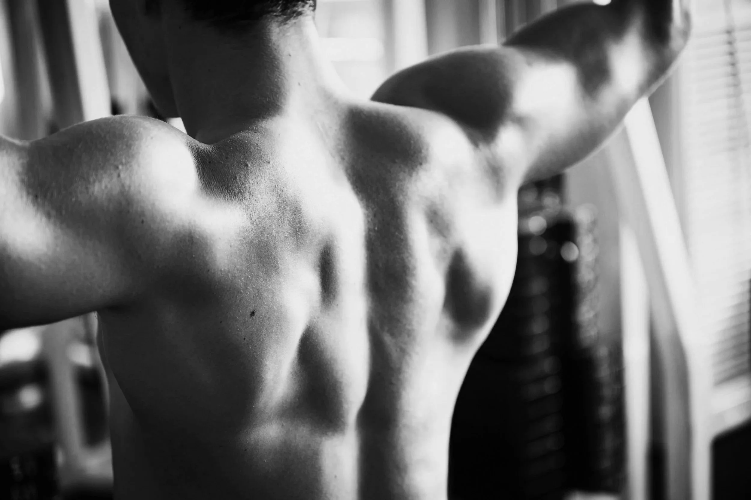Black and white photo of a male torso with defined muscles, arms raised, with blurred background.