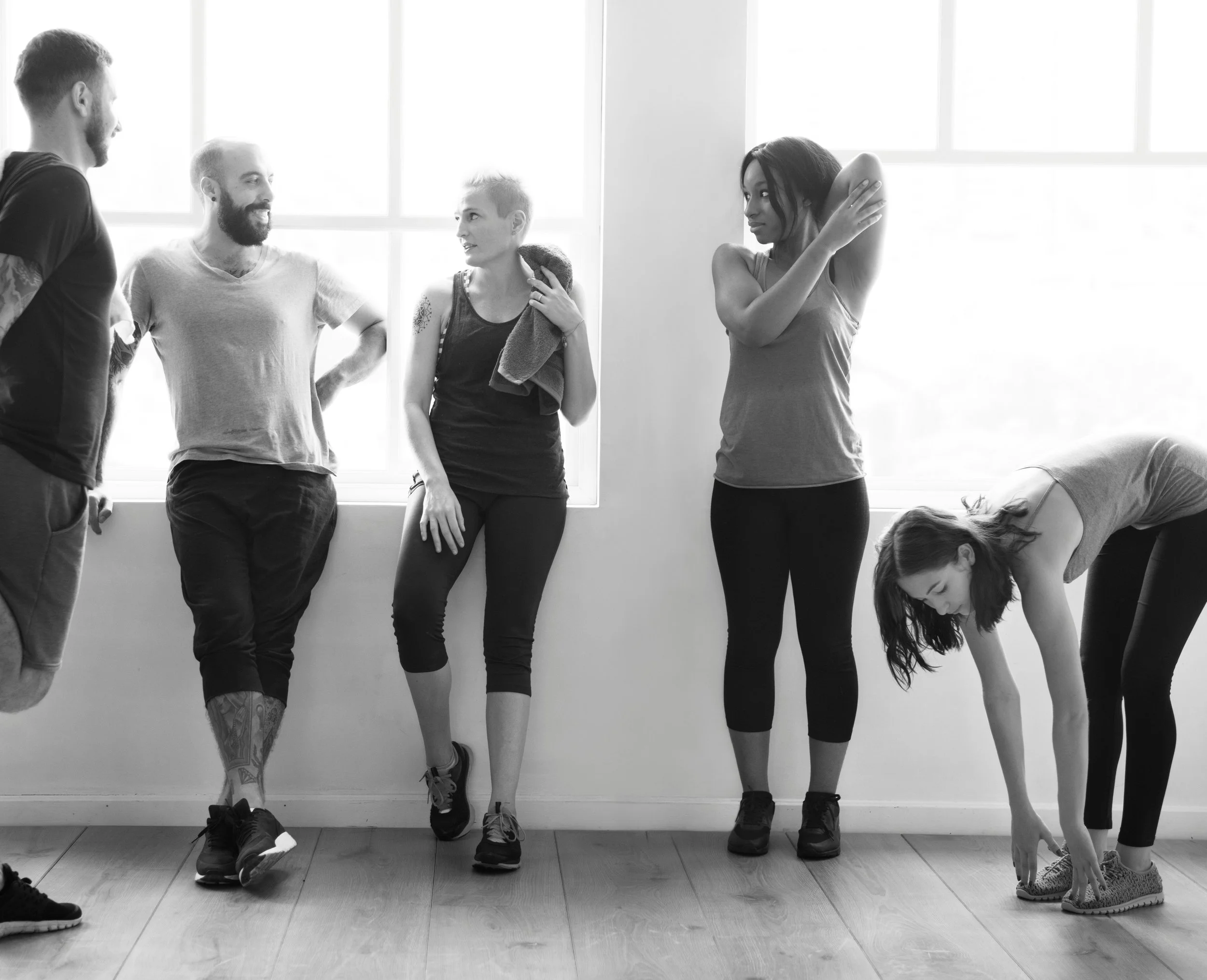 Group of five people in workout gear standing and stretching in a bright room with large windows, some talking and one bending down to touch their toes.