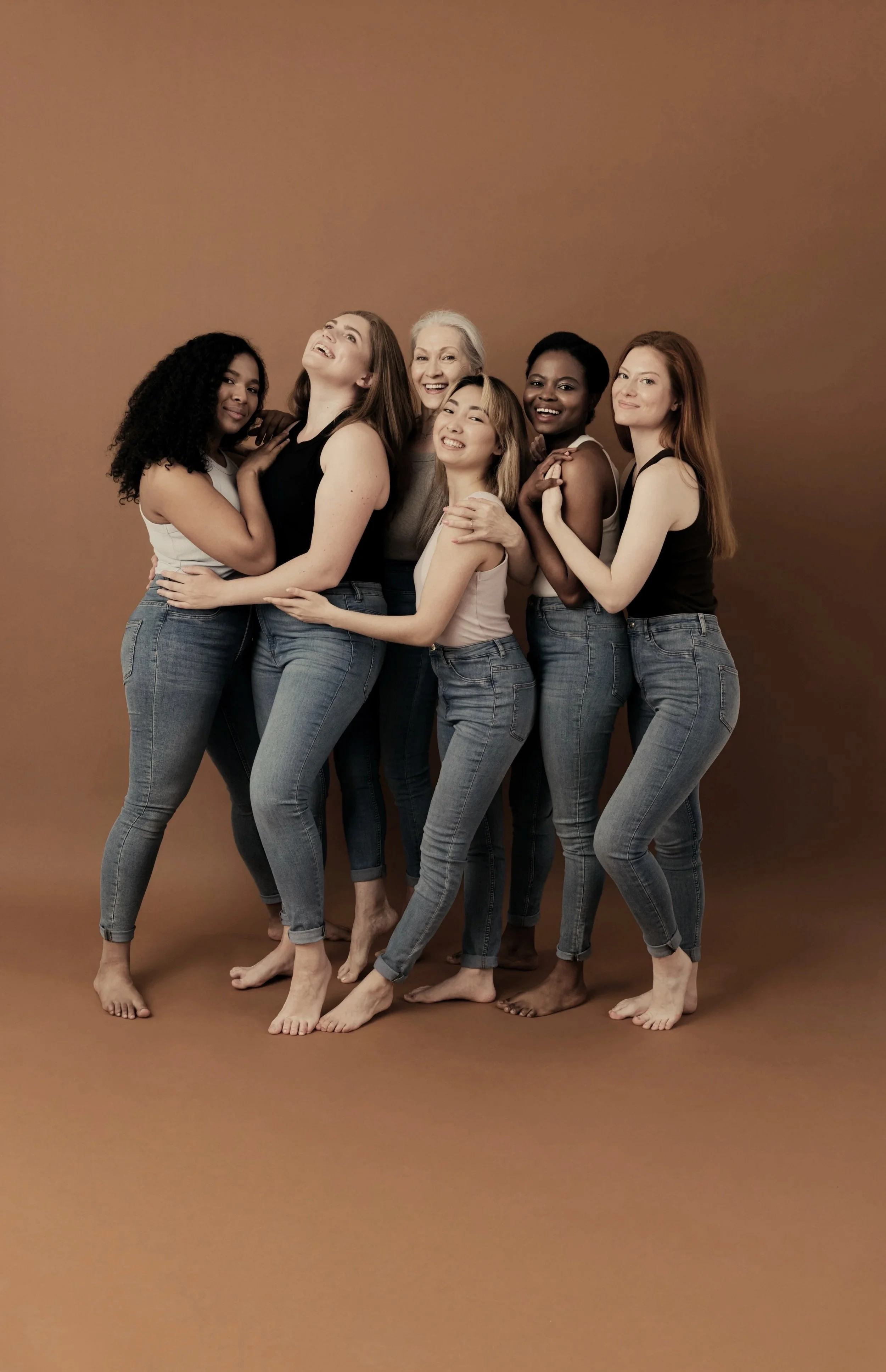 Group of seven women of diverse ages and ethnicities embracing and smiling against a plain brown background.