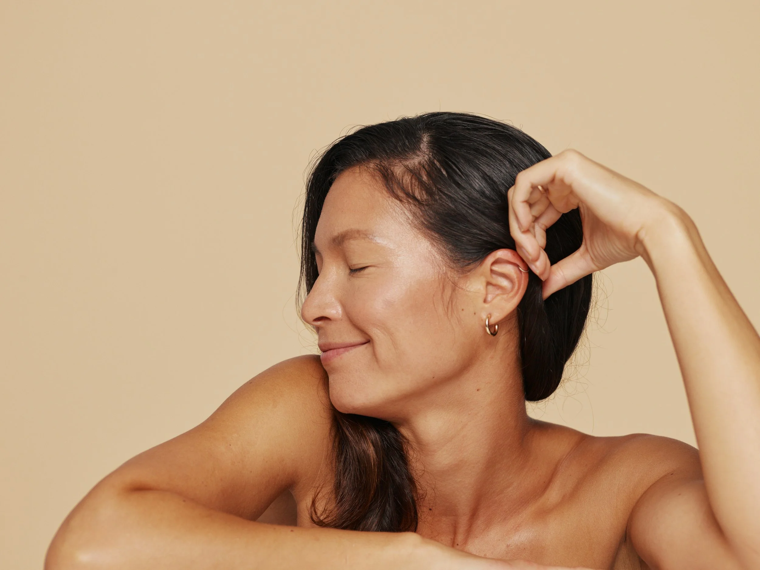 A woman with dark hair and hoop earrings smiling with her eyes closed, resting her arm on a surface, against a beige background.