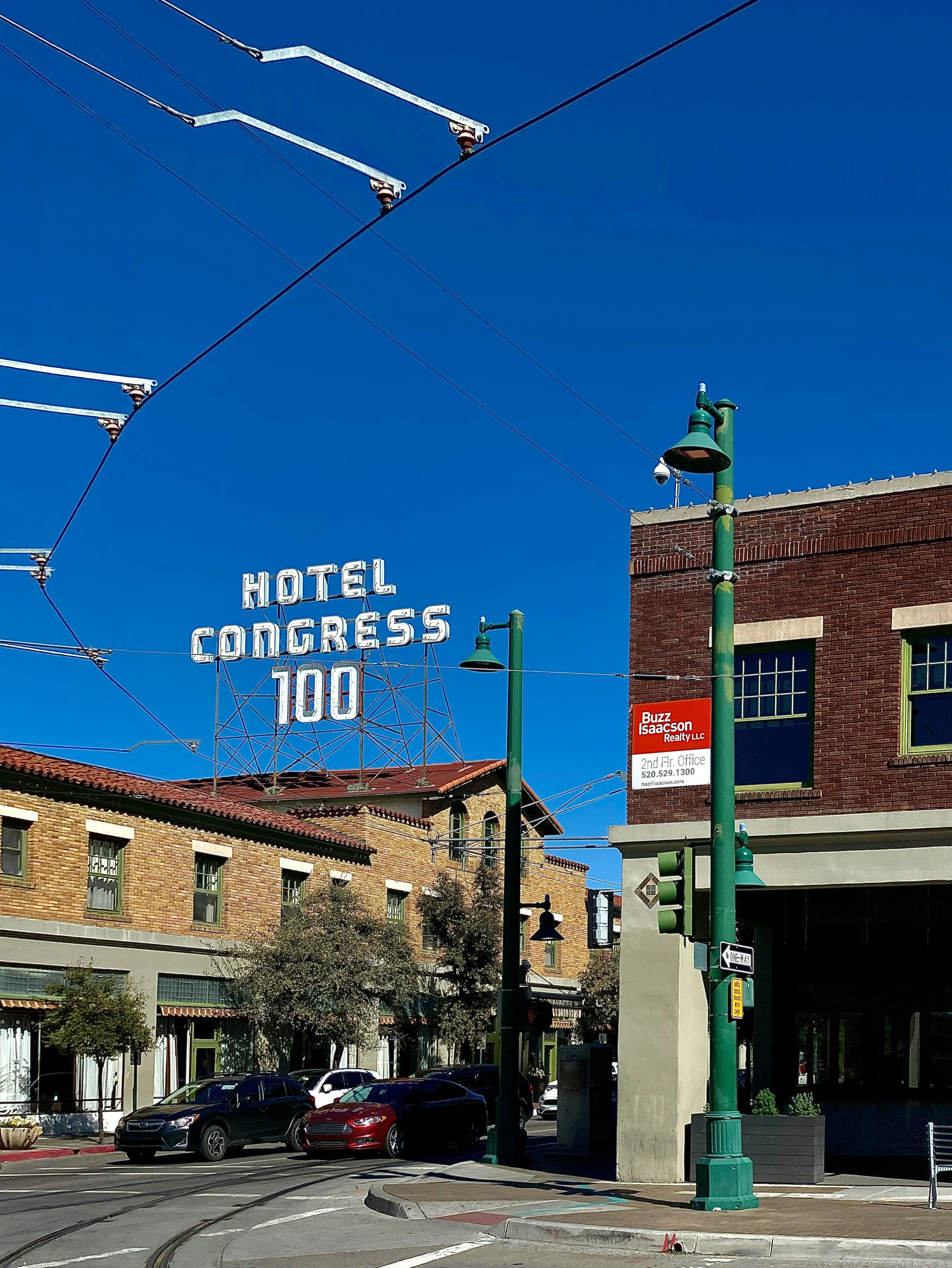 Street view with a large neon sign reading 'Hotel Congress 100' above a building, street cars, cars parked along the street, green streetlights, brick buildings, and a blue sky.
