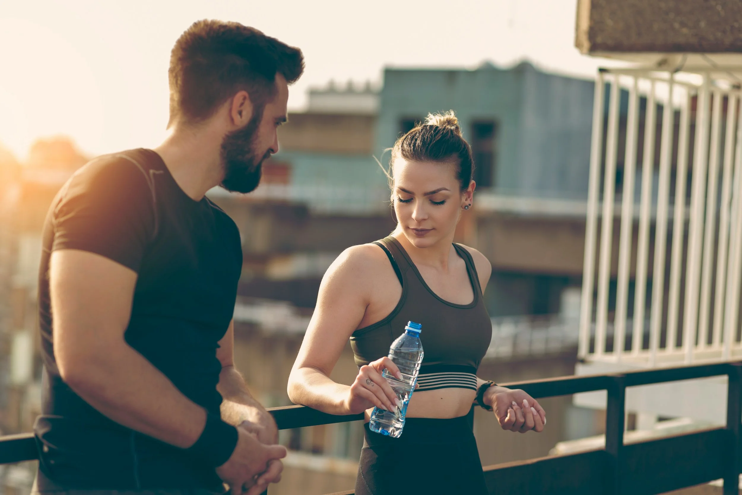 A man and woman in athletic clothing on a rooftop, with the woman holding a water bottle, engaging in conversation during sunset.