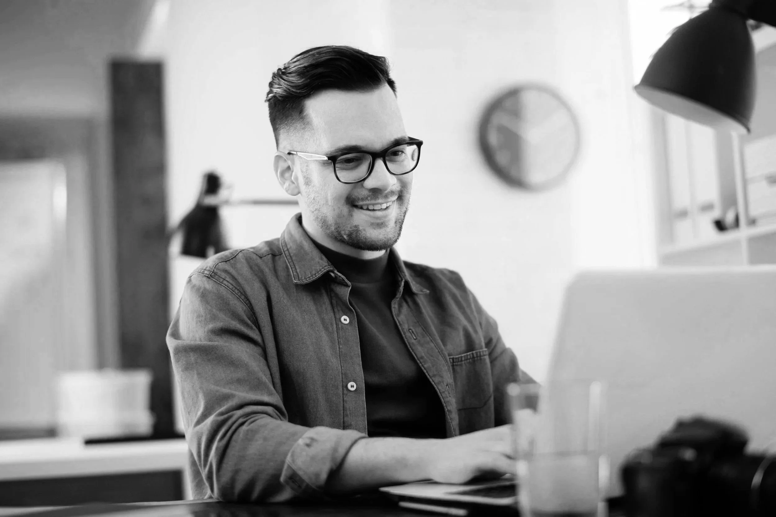 A young man with glasses, smiling, sitting at a desk and working on a laptop in a modern office.