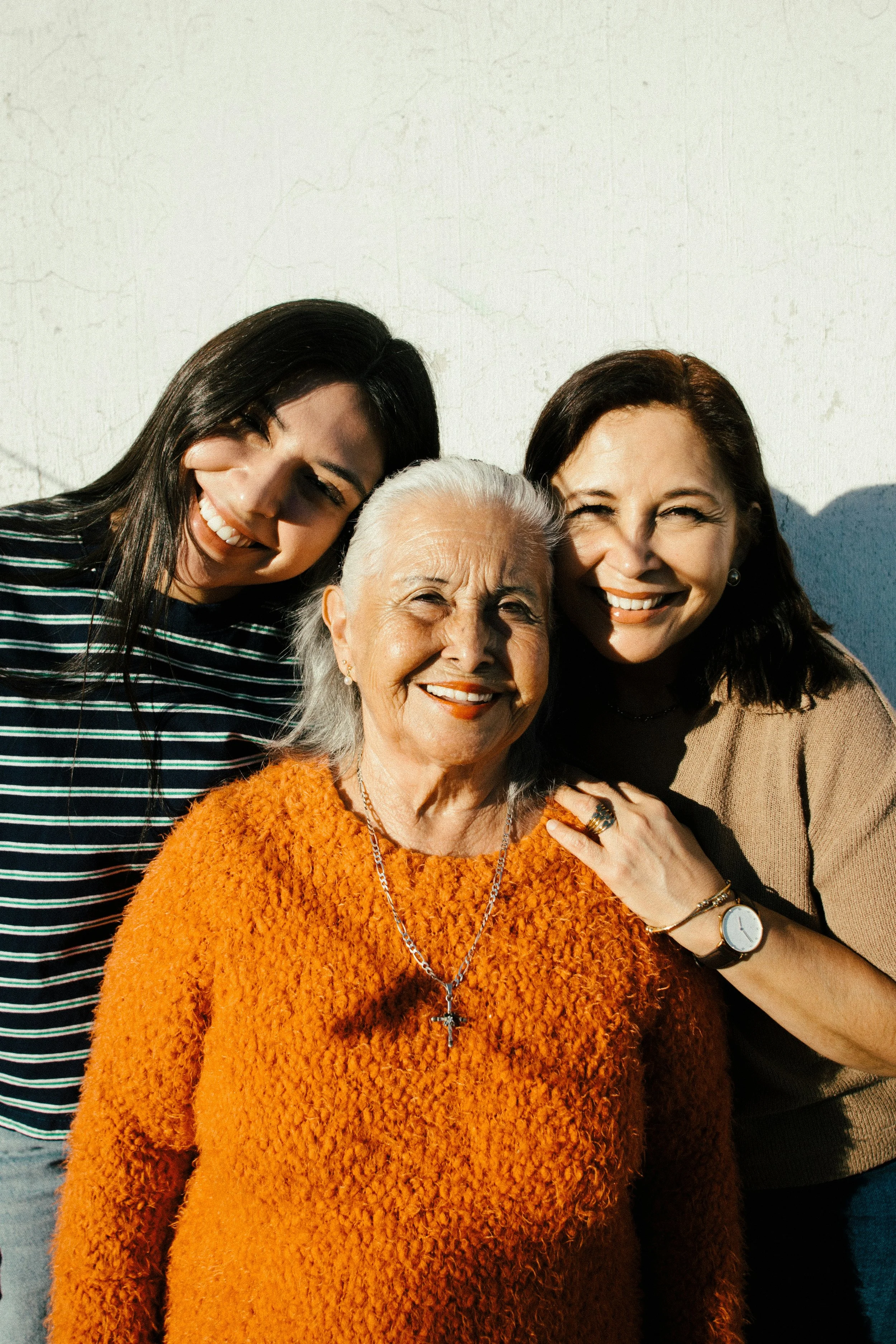 Three women smiling and posing together outside in sunlight, with a white textured wall in the background, including an elderly woman in the center wearing an orange sweater, a silver cross necklace, and a watch, with two younger women on either side.