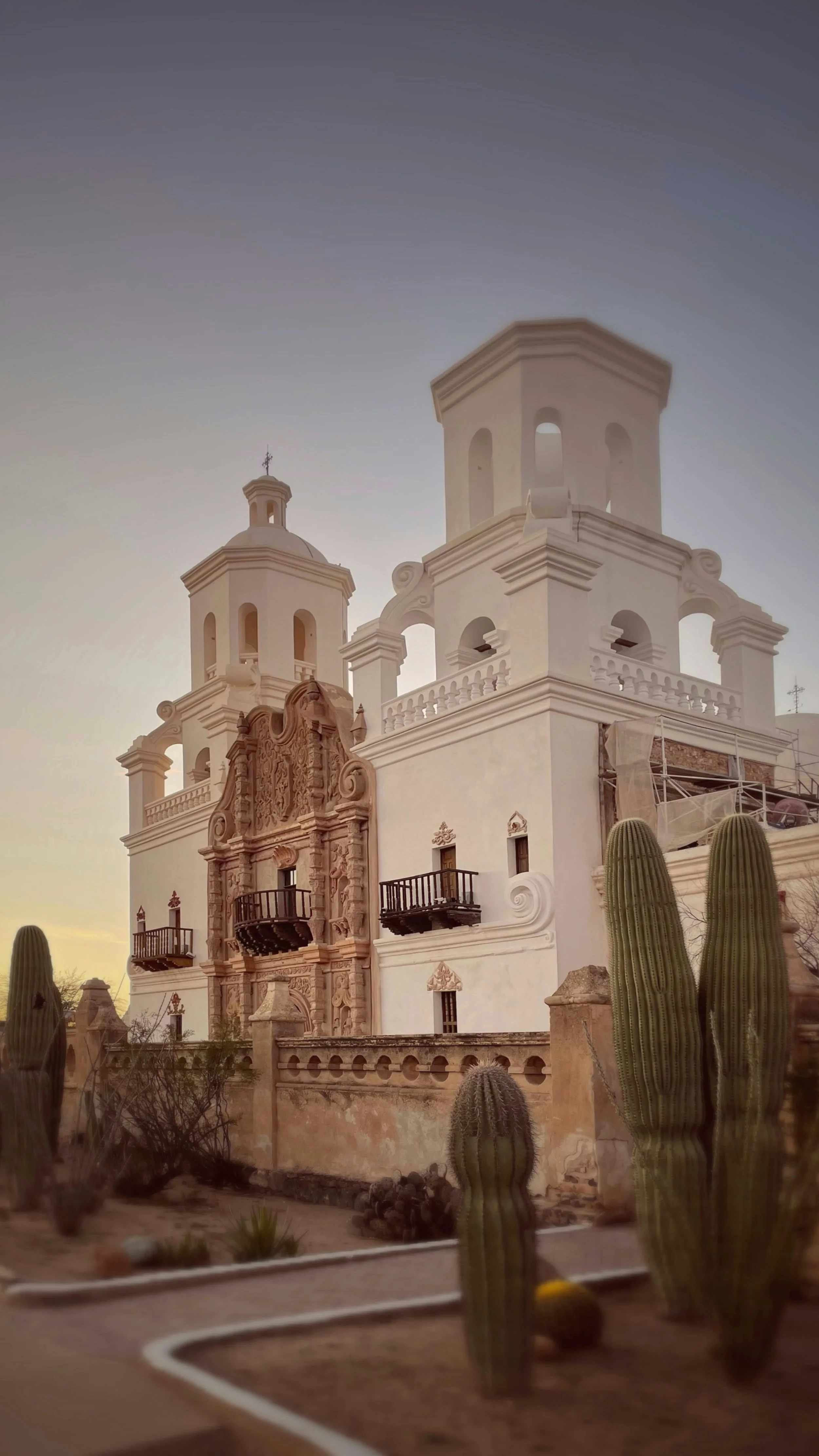 A historic church with intricate architectural details, white walls, and twin bell towers, with desert cacti in the foreground during sunset.