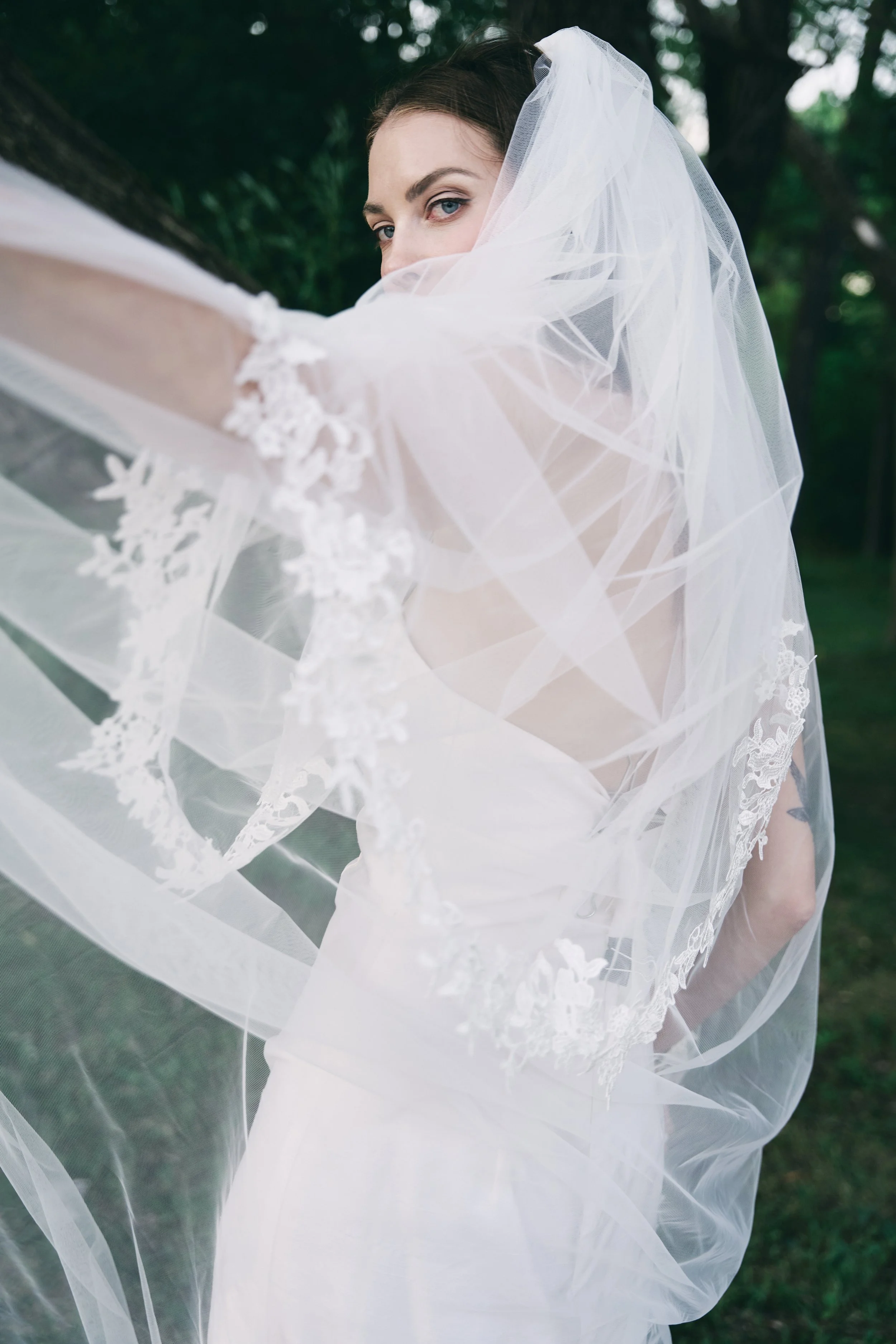 A woman in a wedding dress and veil standing outdoors, partially obscured by her flowing veil, with trees in the background.