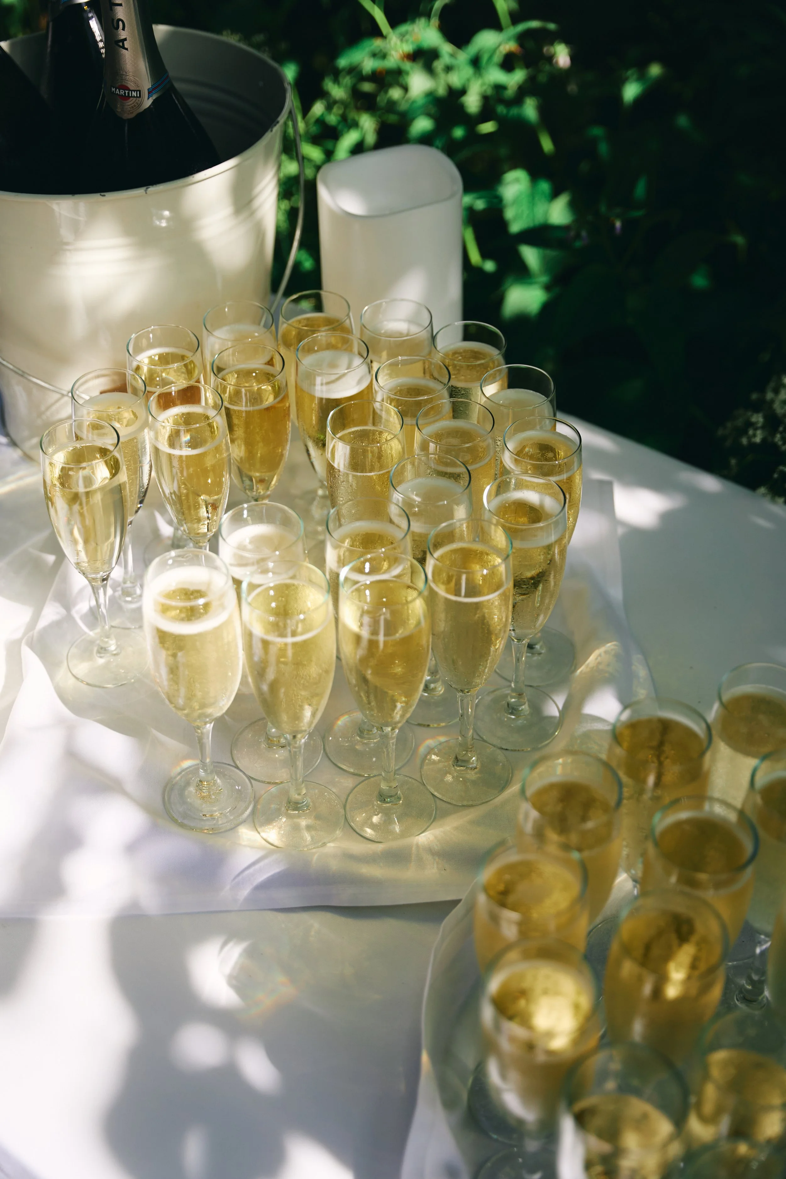 Multiple glasses of champagne on a white table, with a bucket of champagne bottles in the background, outdoors with greenery.