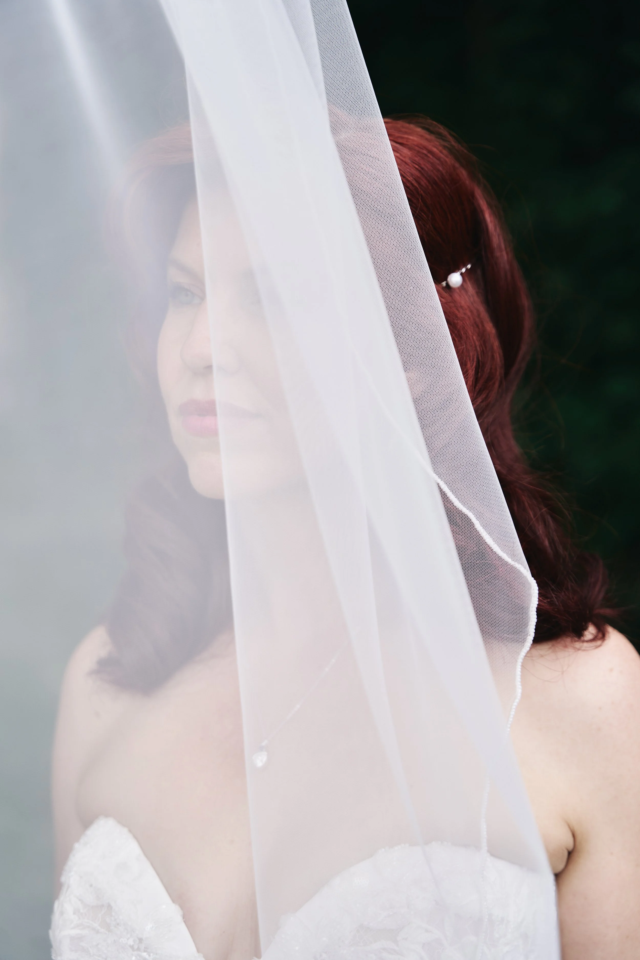 A woman with red hair in a wedding dress, partially obscured by a sheer white veil, outdoors with a dark green background.