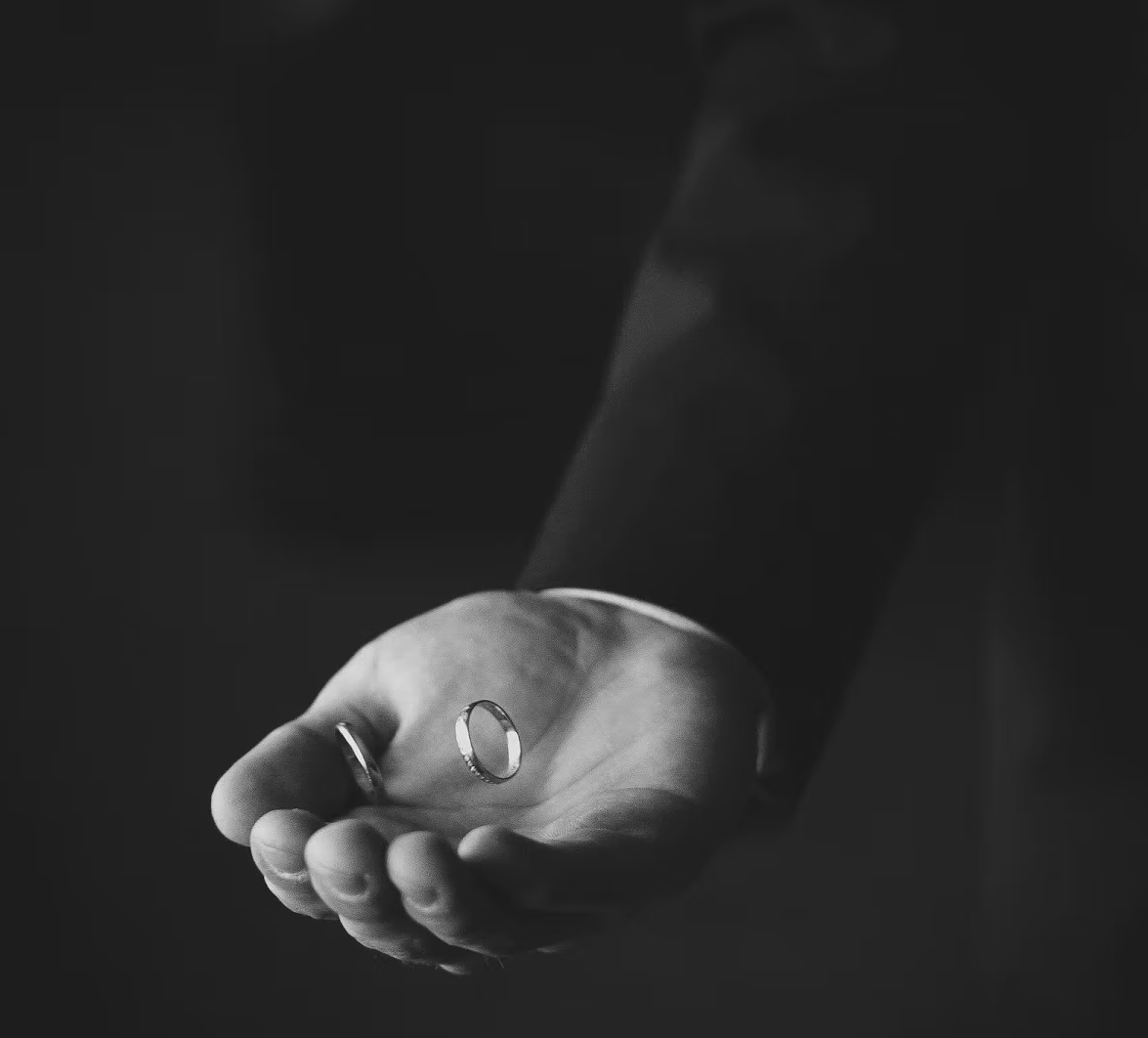 A person's hand holding two wedding rings, one ring on the middle finger and the other on the ring finger, in black and white.