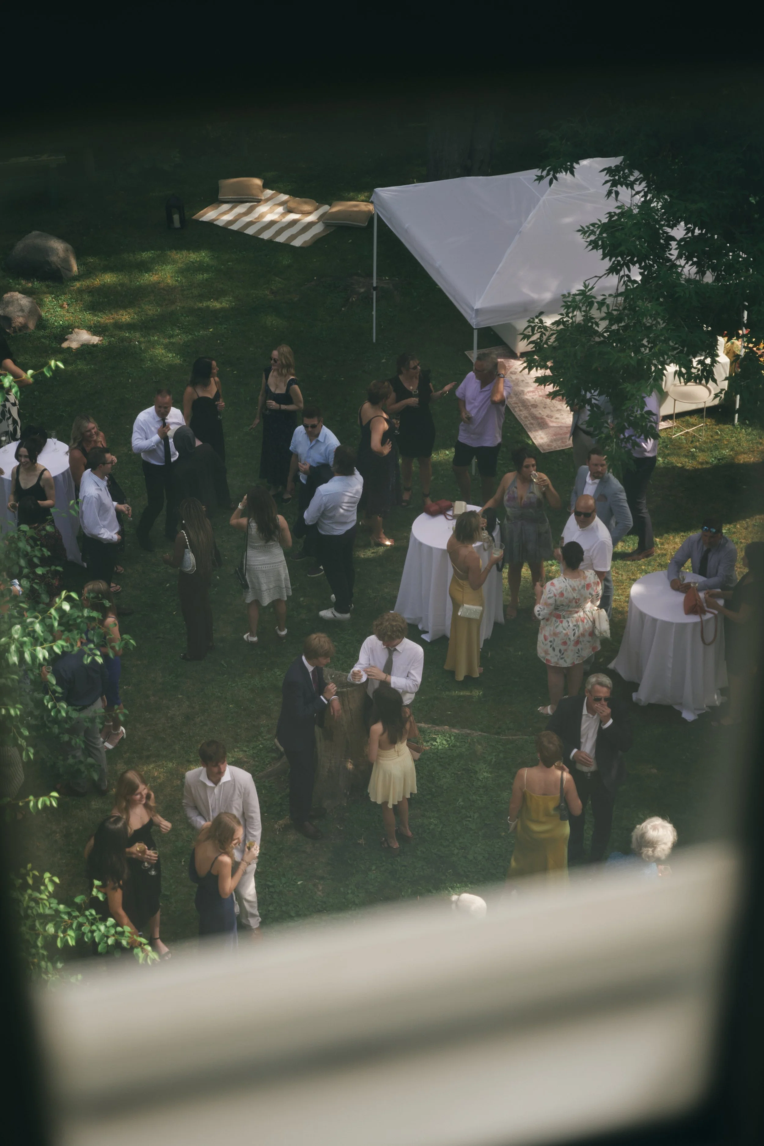 People gathered outdoors at a social event or party on a lawn, with tables, lounge chairs, and a white canopy.