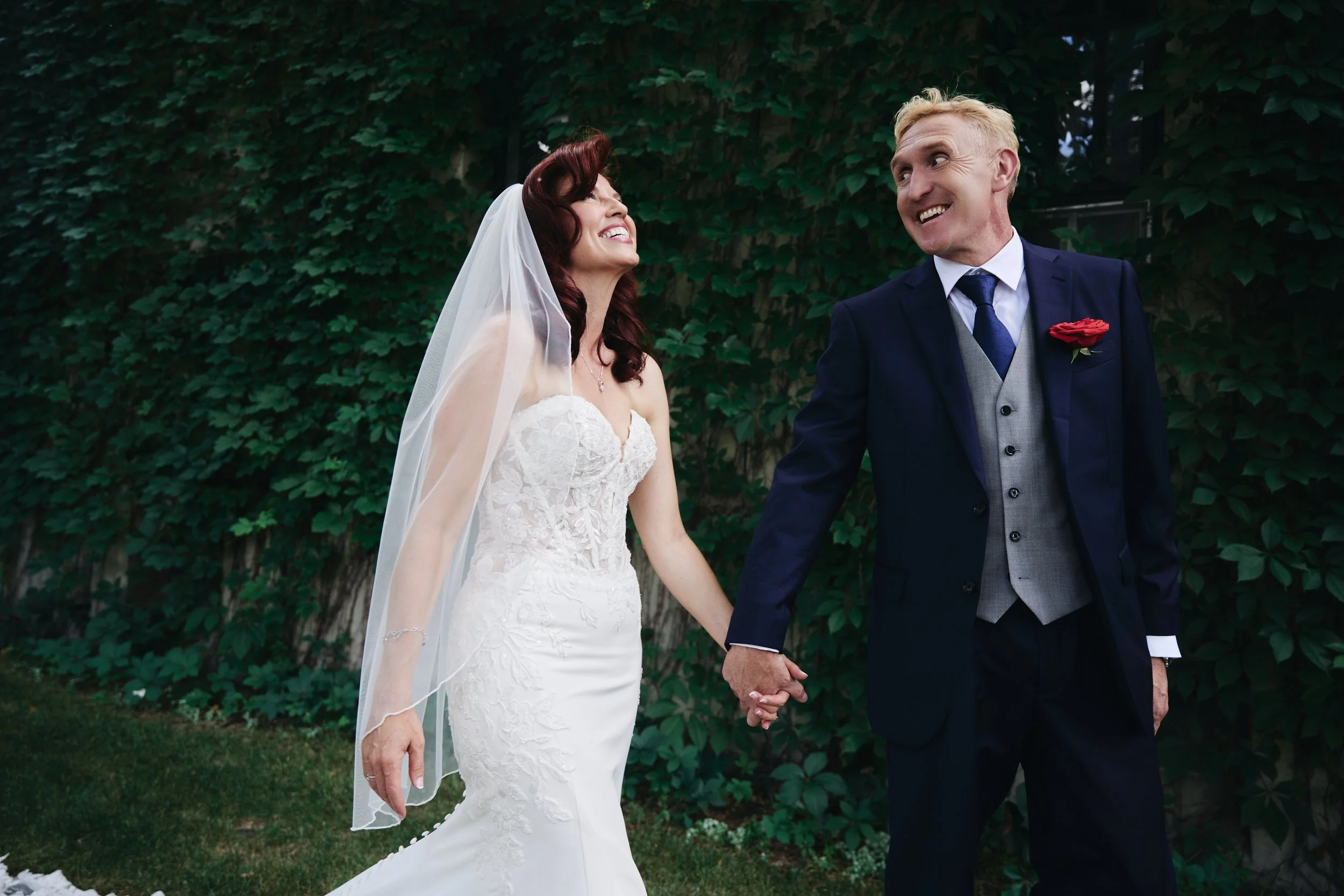 A newlywed couple holding hands and smiling outdoors with a leafy green background.