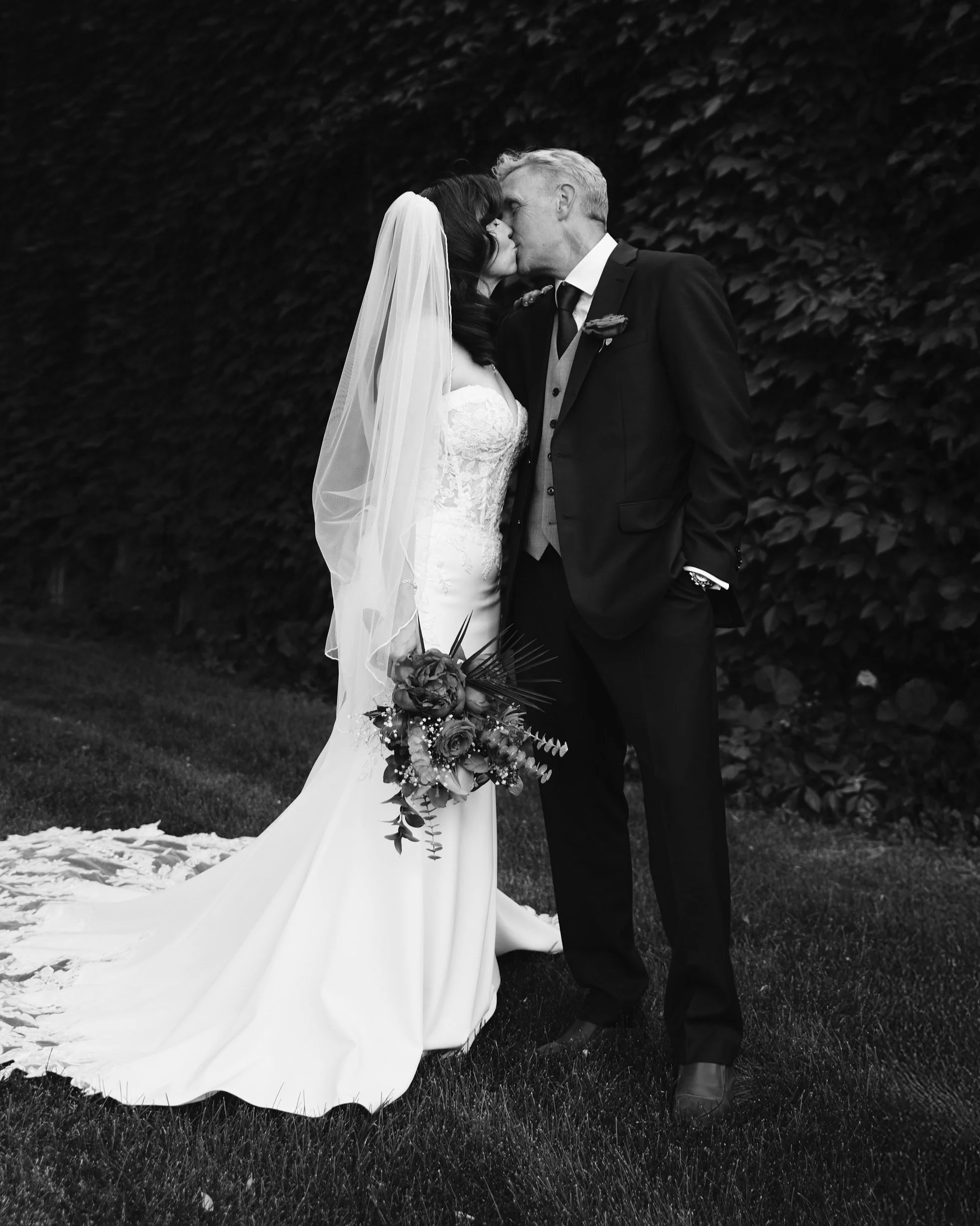 Black and white photo of a bride and an older man, likely her father, sharing a kiss at a wedding. The bride is in a strapless wedding gown, holding a bouquet, with a veil over her dark hair. The man is in a dark suit with a boutonniere, standing out