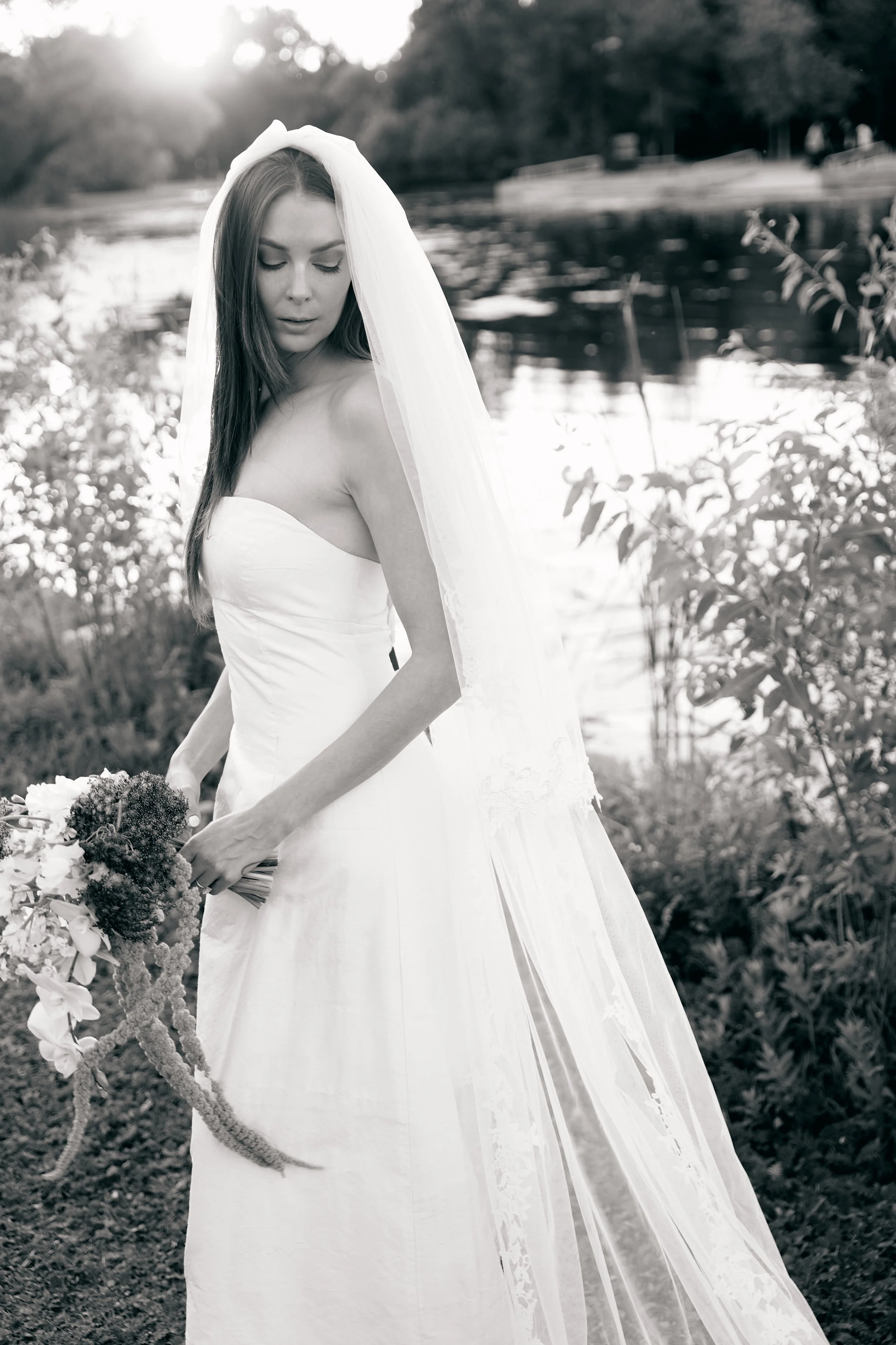 A woman in a wedding dress holding a bouquet standing outdoors near a body of water with trees in the background.