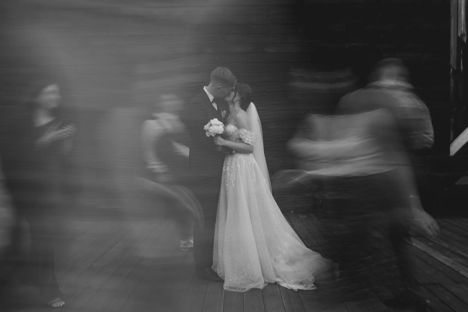 Black and white photo of a bride and groom dancing closely at a wedding reception, with blurred guests dancing around them.