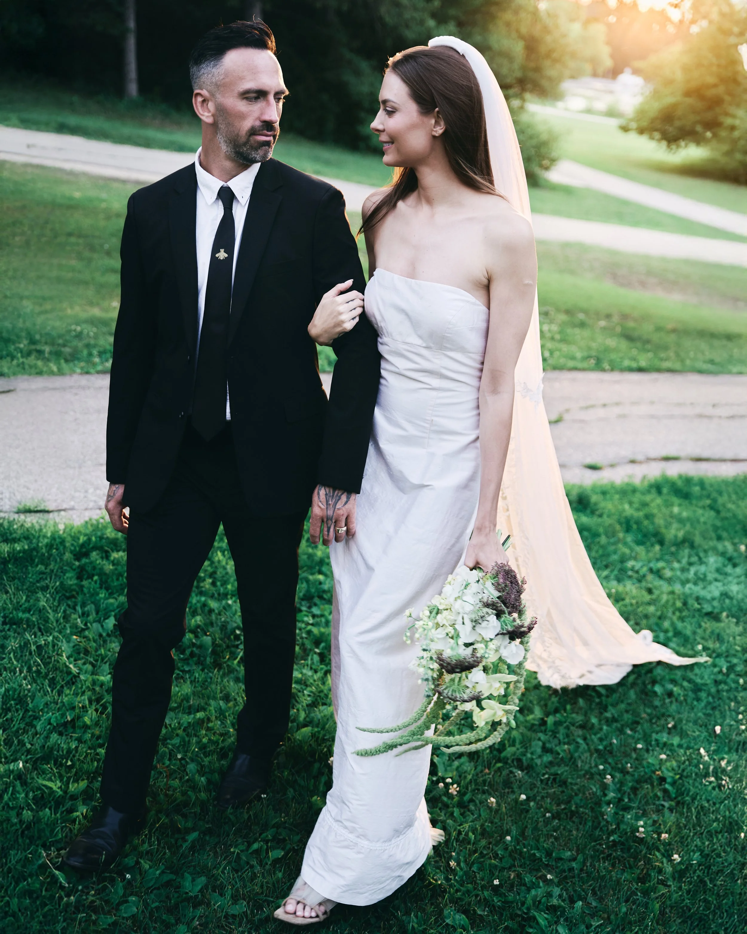 A man in a black suit and woman in a strapless white wedding dress walking together outdoors on a sunny day, with the woman holding a bouquet of flowers.