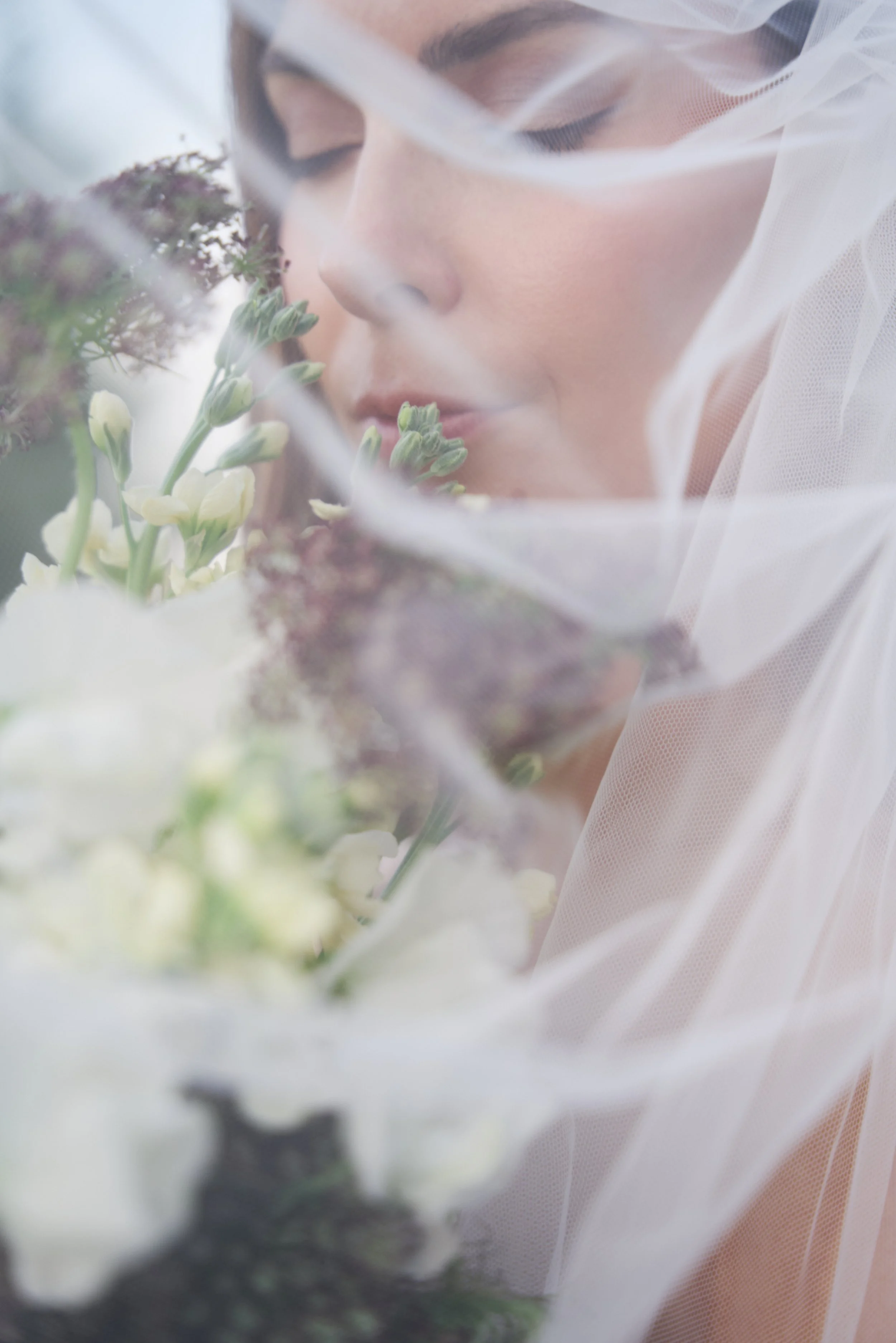 Close-up of a woman with a veil and makeup, gently smelling a bouquet of white and purple flowers.