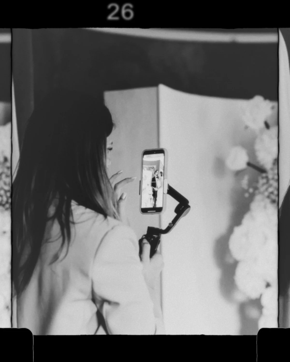 A woman taking a photo with a smartphone mounted on a stabilizer, capturing her reflection in a mirror surrounded by floral decorations, in black and white.