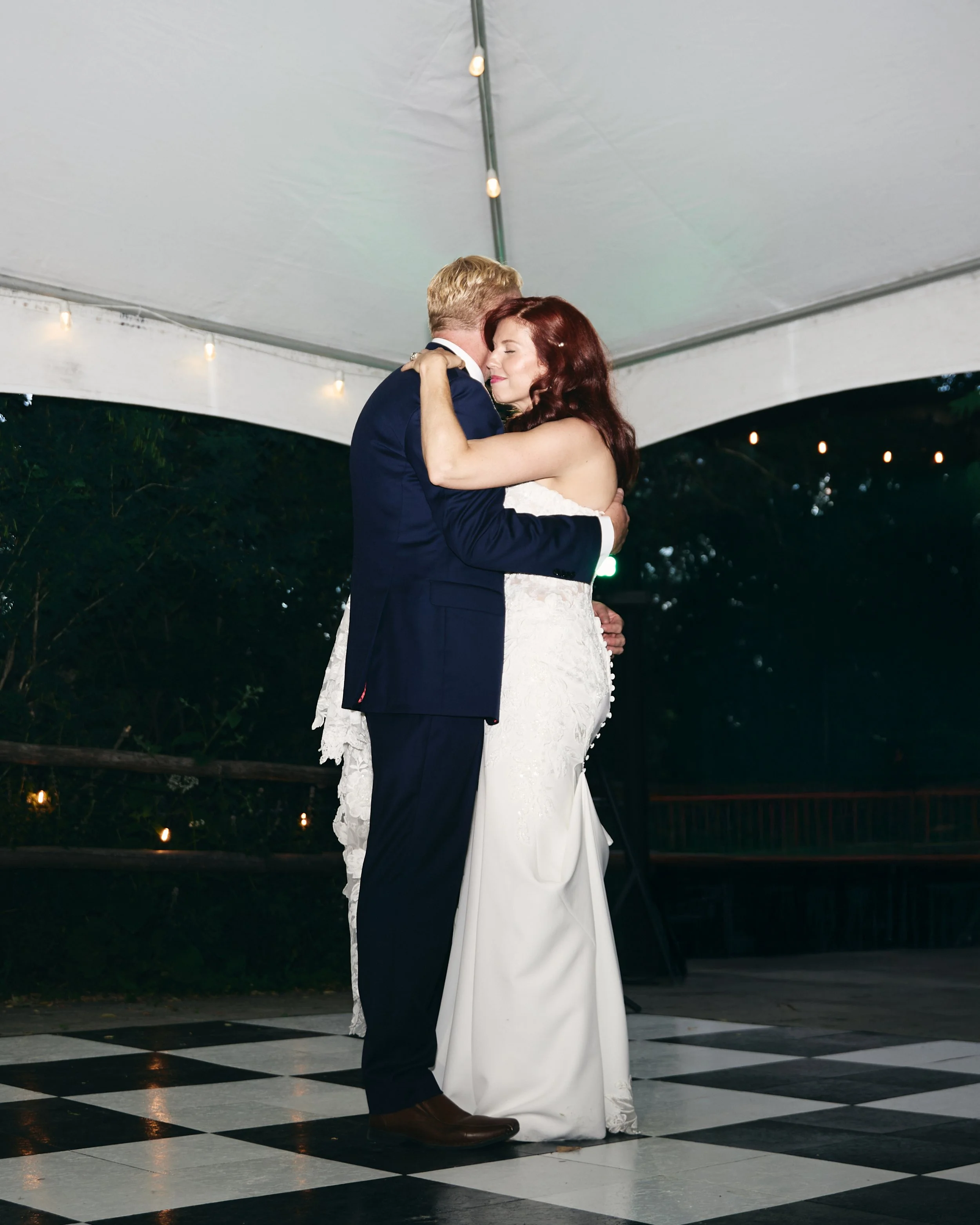 A bride and groom are hugging during their wedding reception under a tent with string lights, on a black and white checkered dance floor.