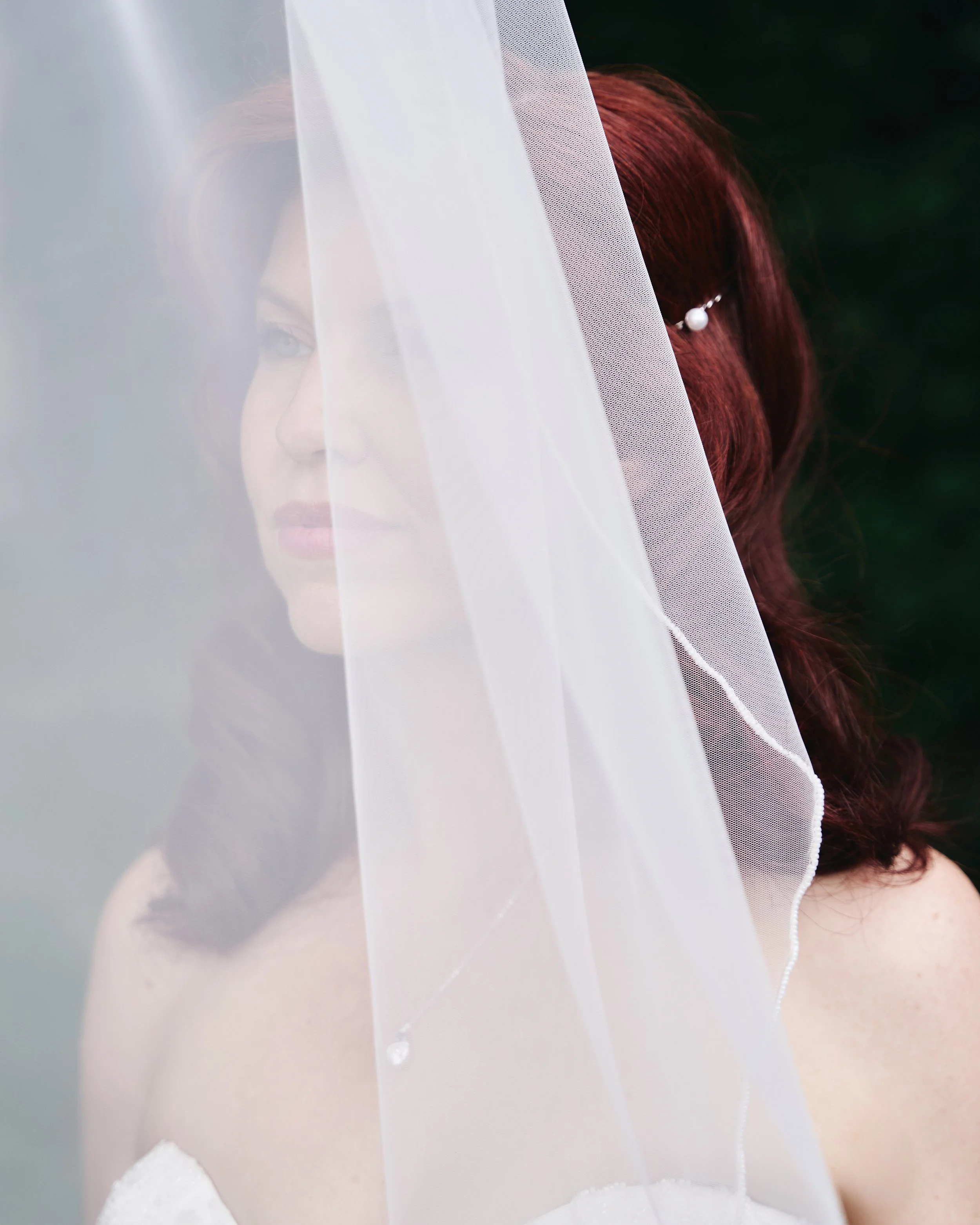 A woman with red hair wearing a white dress and pearl jewelry nose behind a sheer veil.