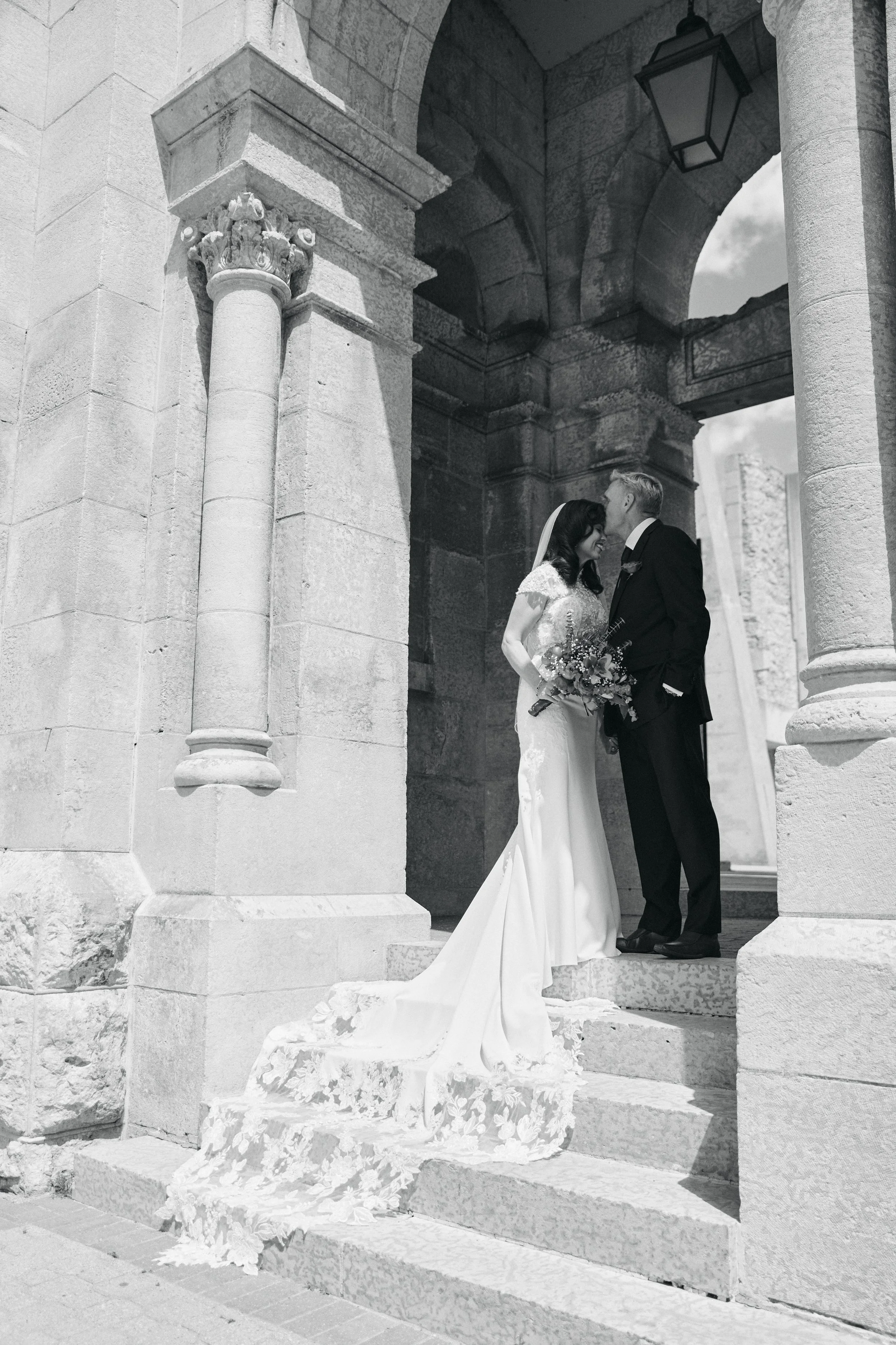 Black and white photo of a bride and groom standing on stone steps under an arch, sharing an intimate moment with their foreheads touching. The bride is holding a bouquet and wearing a wedding dress with a lace train. The groom is in a suit.