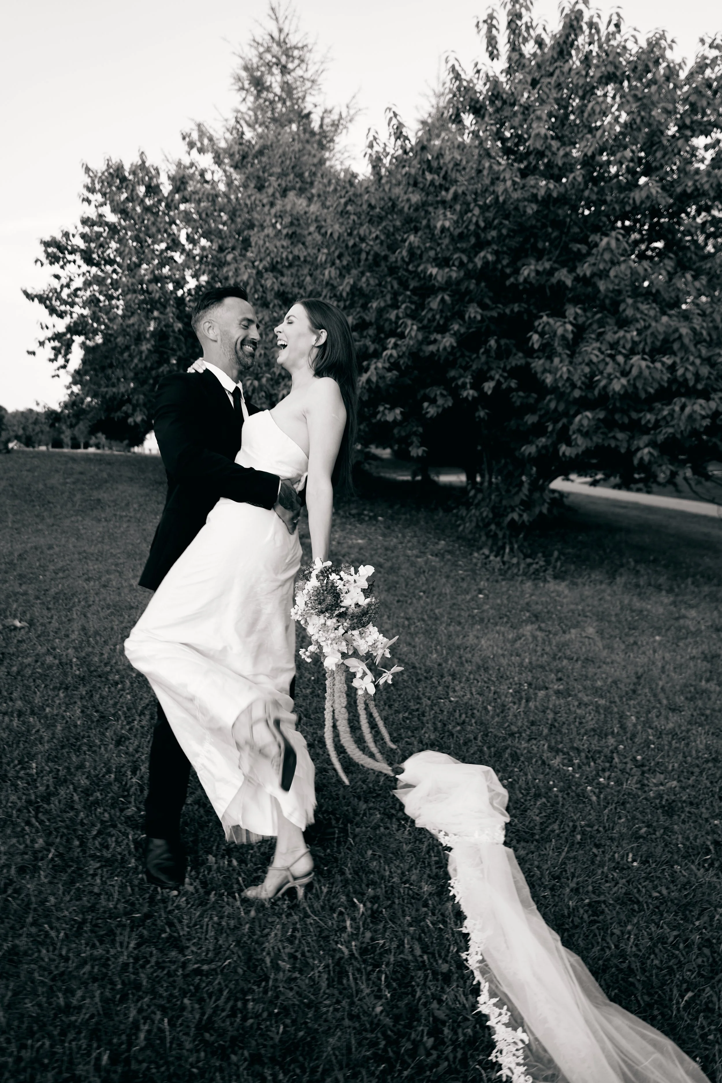 A black and white photo of a newlywed couple outdoors, with the groom lifting the bride and both laughing. The bride is holding a bouquet, and a long veil trails on the grass. Large trees are in the background.