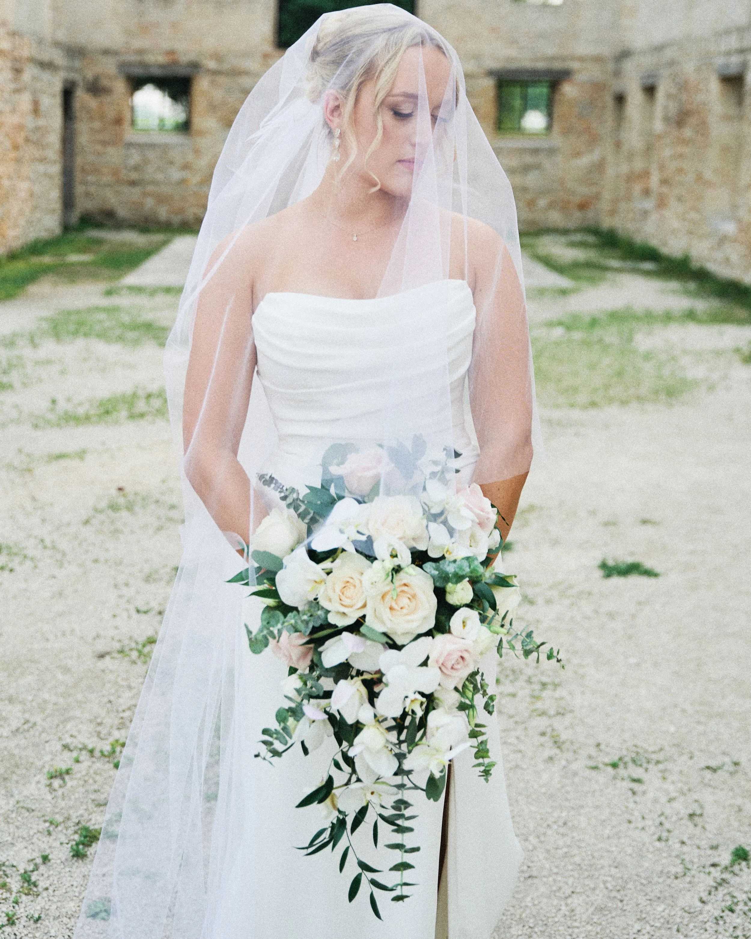 Bride in a white strapless wedding dress holding a cascading bouquet of white and blush roses, white orchids, and greenery, standing outdoors with old stone ruins in the background, wearing a veil and pearl earrings.