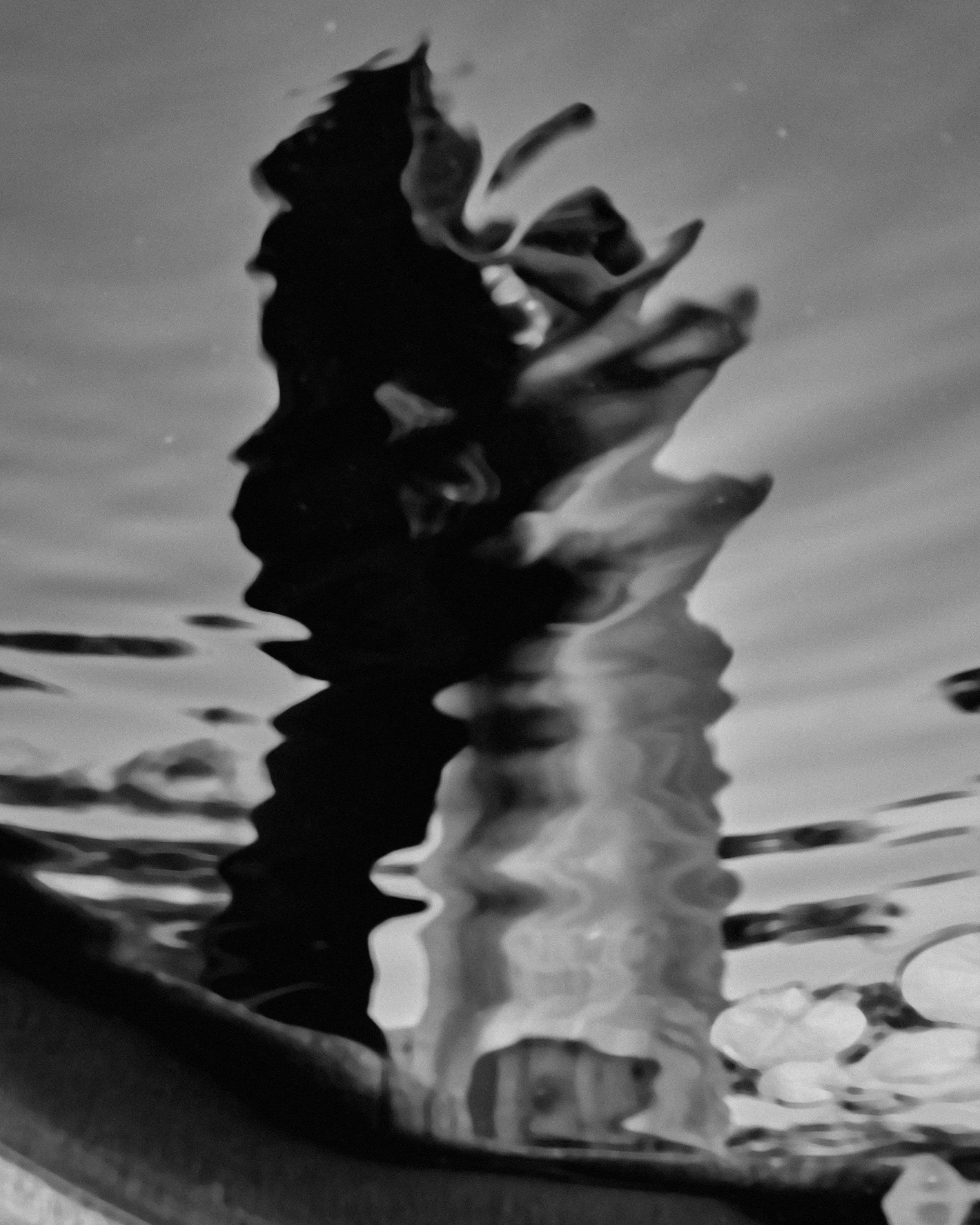 Black and white photo of a loosely folded or crumpled paper submerged in water, with reflections and ripples on the water's surface.