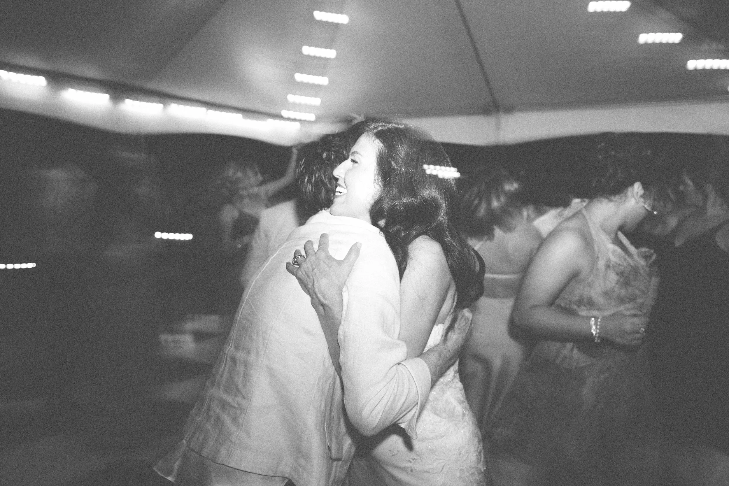 A black and white photo of a woman and man dancing closely, smiling and embracing, at a dance event or celebration with other women dancing in the background.