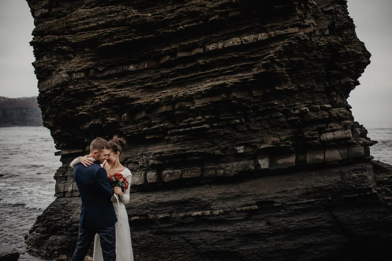 A bride and groom standing close together on a rocky shoreline, with the bride holding a bouquet of red and white flowers, and a large dark cliff behind them on a cloudy day.