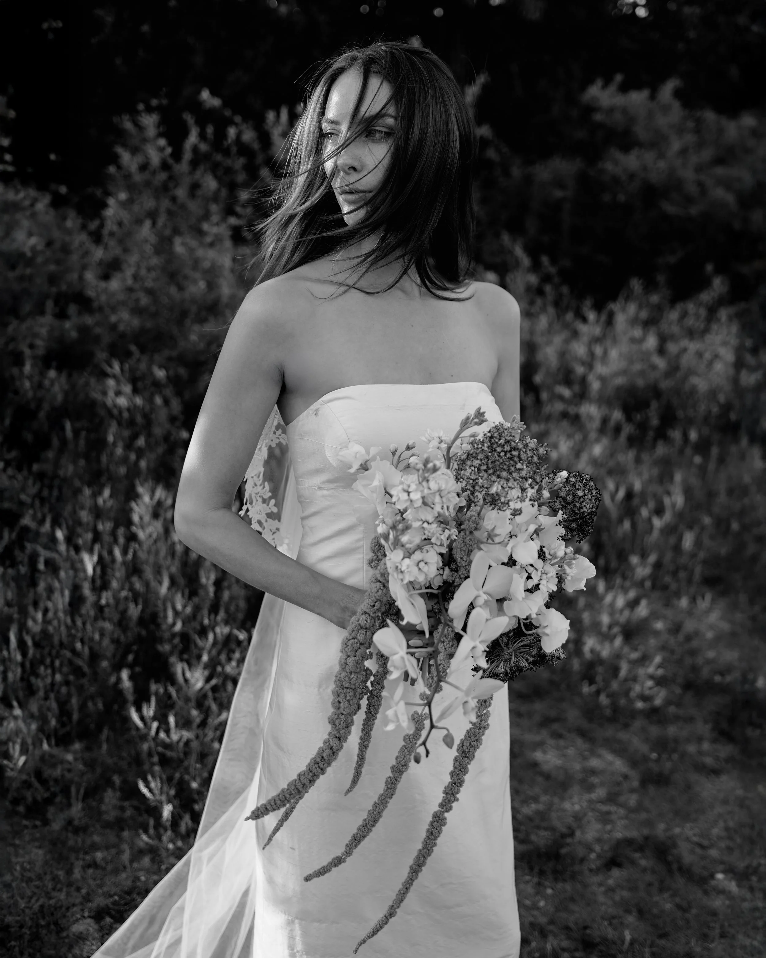 A woman in a strapless wedding dress holding a large bouquet of flowers outdoors.