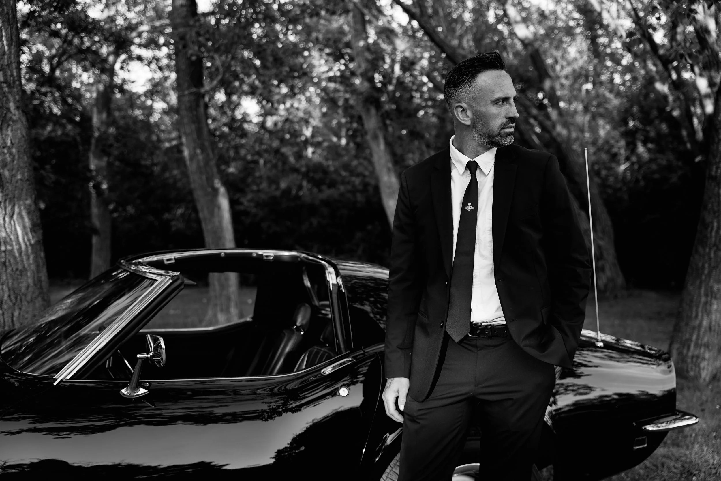 A man in a suit standing next to a vintage convertible car on a lawn with trees in the background.