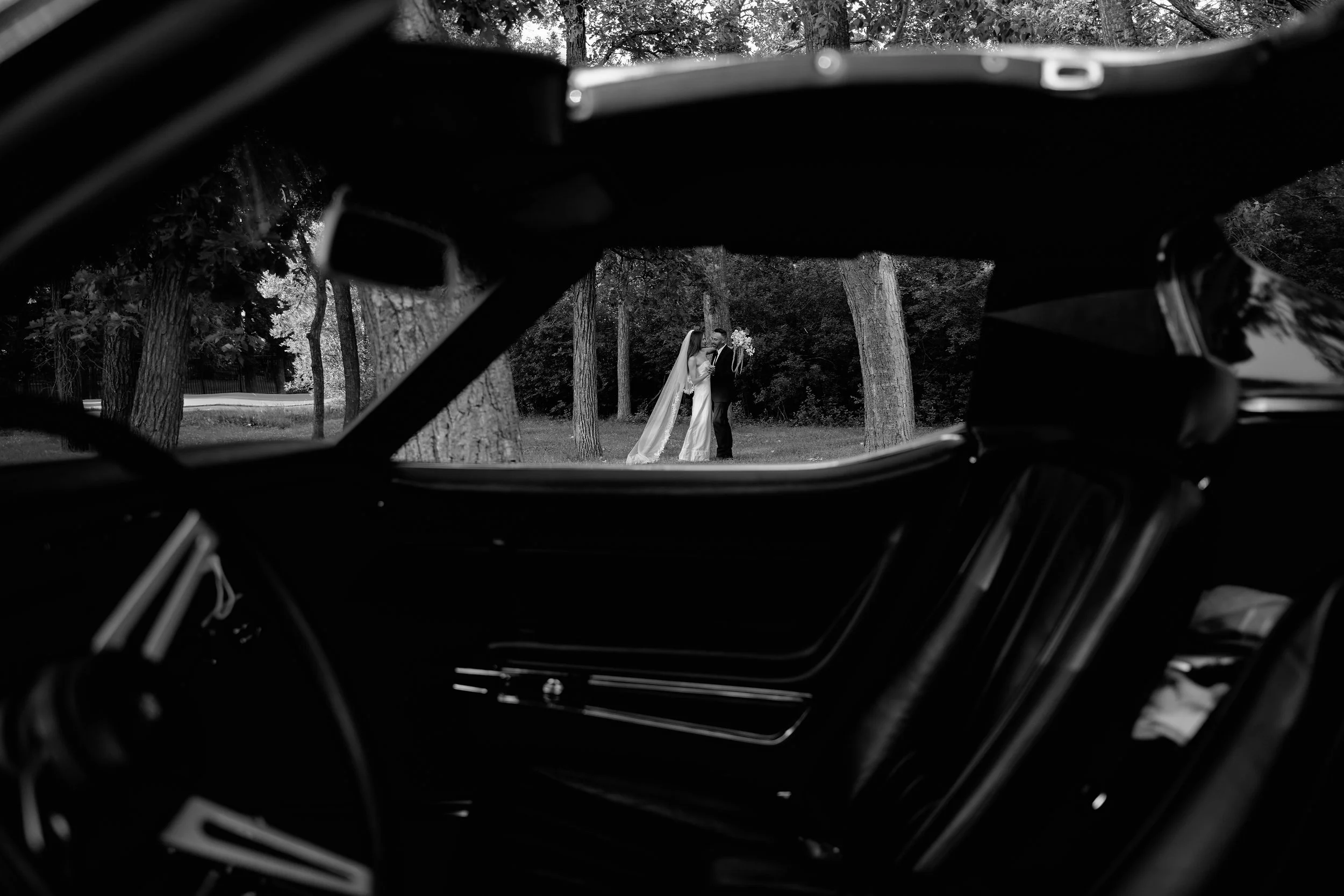 A black and white photo of a bride and groom kissing in a wooded park, seen through the open back of a classic car.