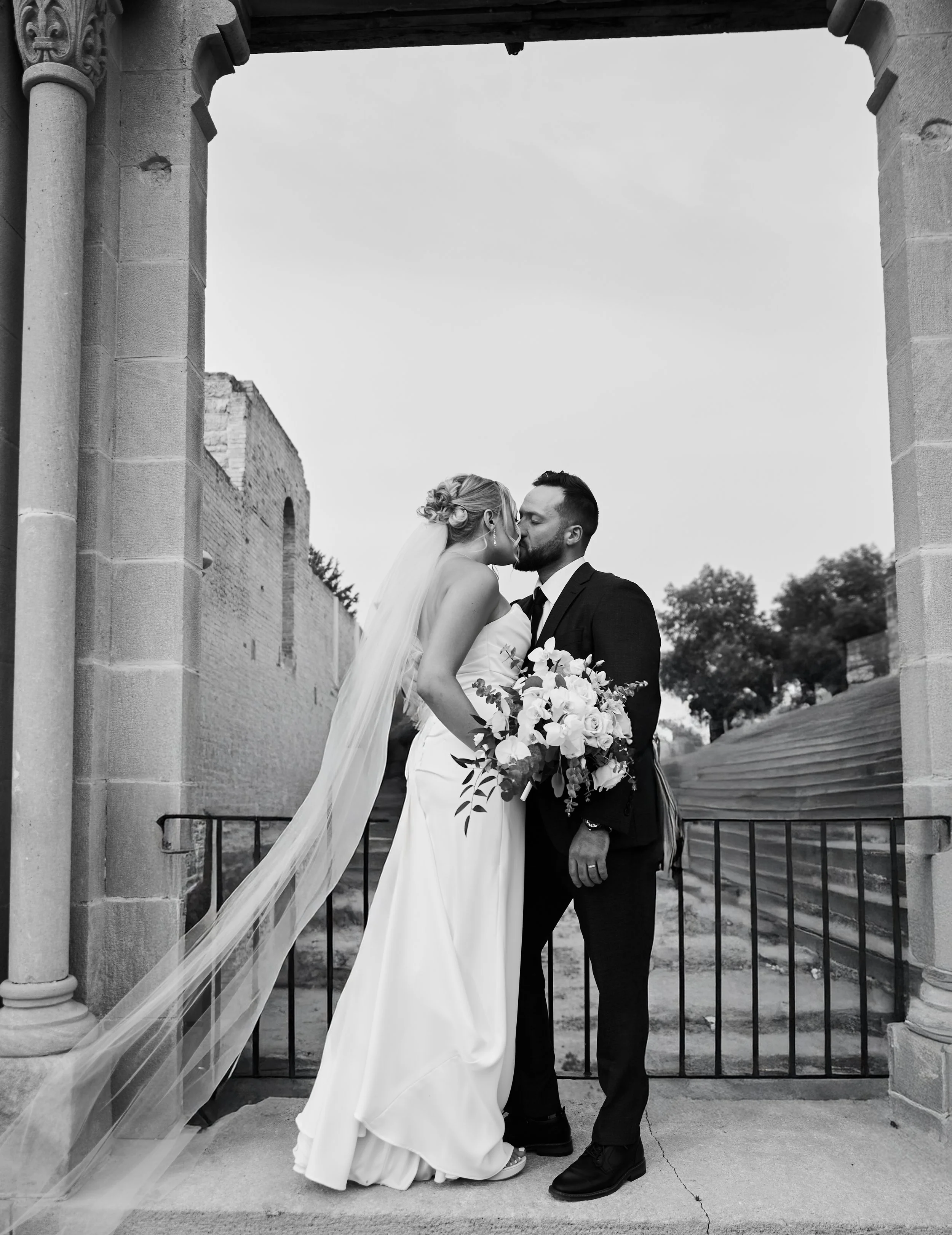 Black and white photograph of a bride and groom kissing on a stone balcony with an archway, holding a bouquet of flowers, during a wedding ceremony.