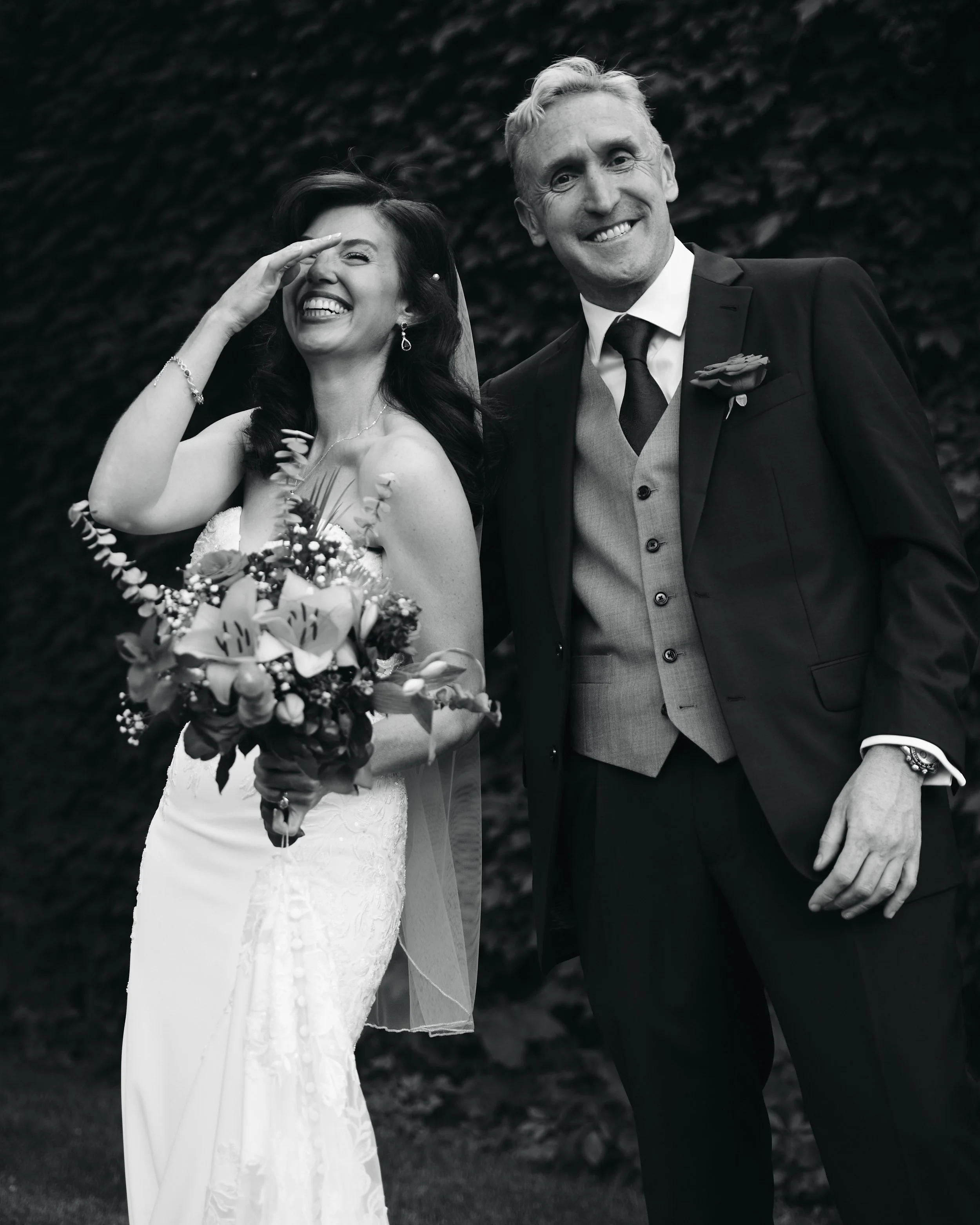 A black and white photo of a bride and an older man (possibly her father) smiling and posing together outdoors, with the bride holding a bouquet of flowers and wearing a wedding dress.
