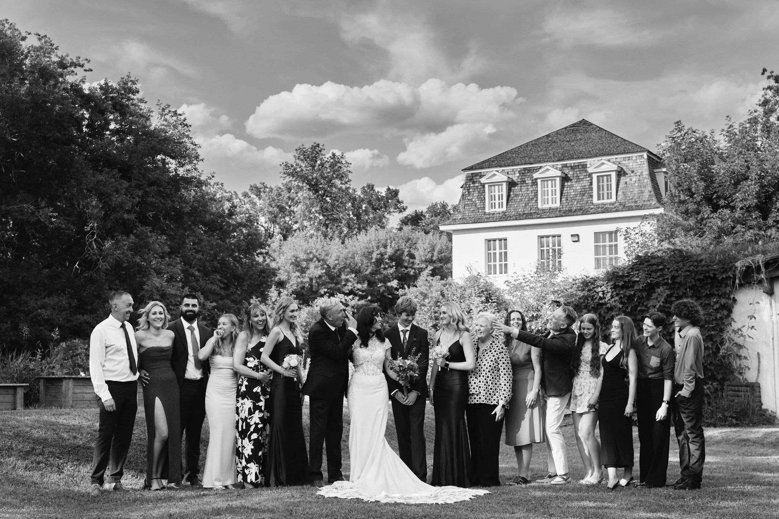 Group of people at a wedding outdoors, with a bride in a wedding dress, groom, and guests, standing on a lawn with trees and a large house in the background.