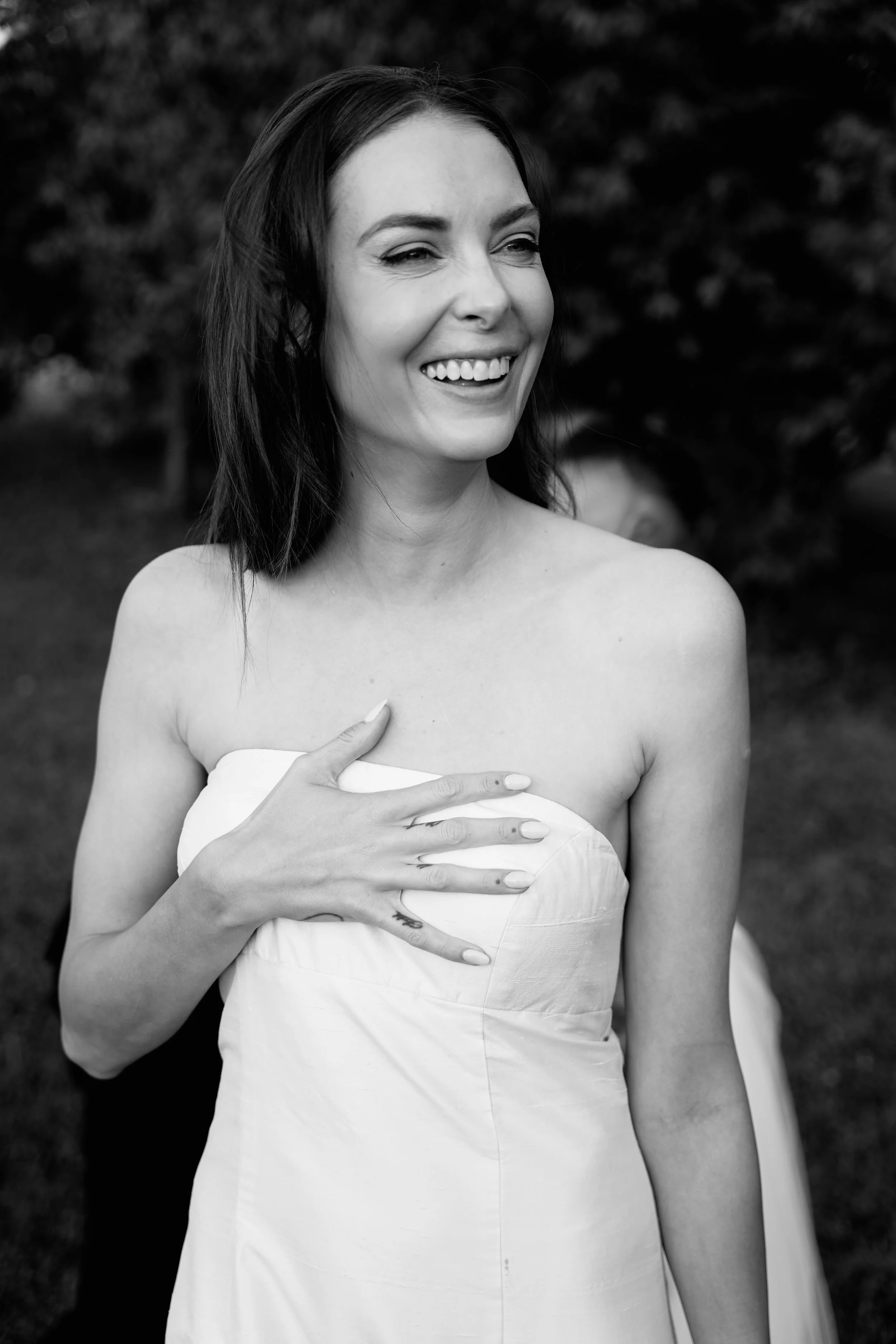 A woman in a strapless dress smiling and covering her chest with her hand outdoors.